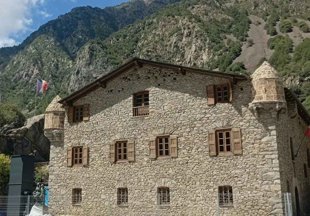 A stone building with wooden shutters and a corner turret, the Casa de la Vall, against a steep forested mountain slope in Andorra la Vella.