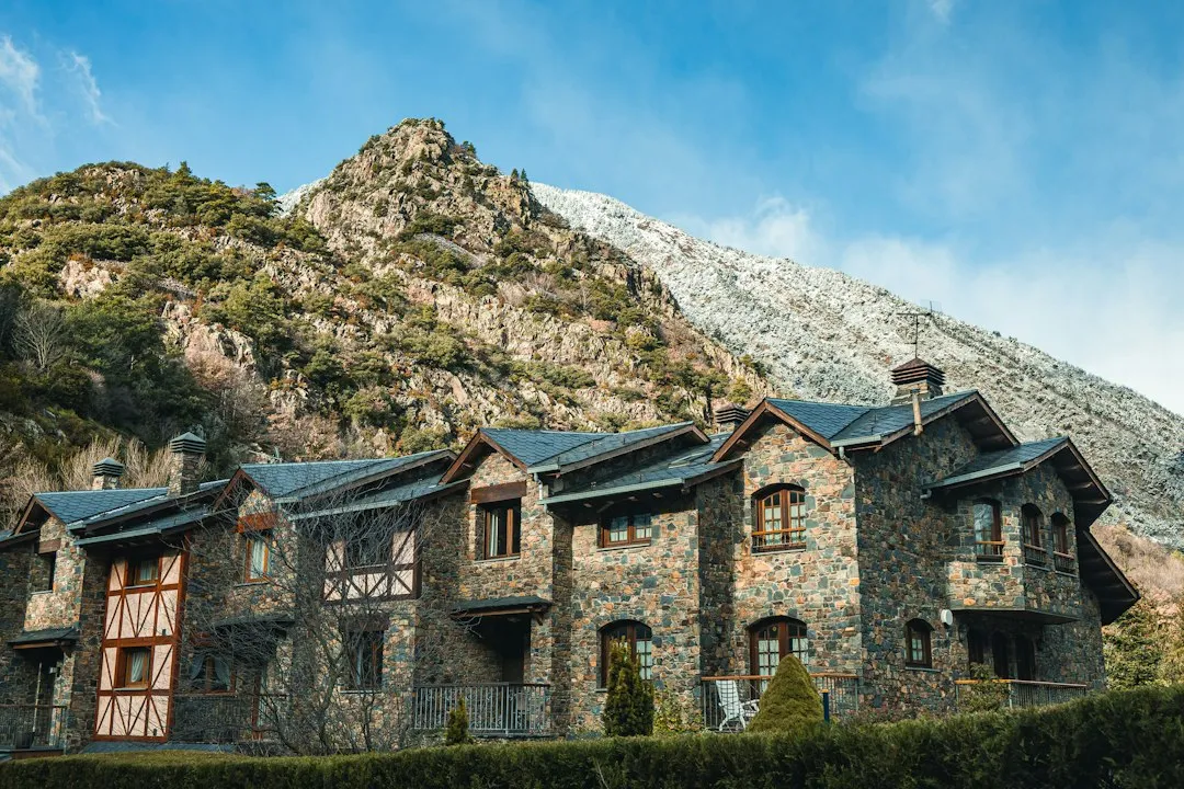 A row of stone chalets with slate roofs and wooden balconies against a steep, bare Pyrenean slope under a blue sky.