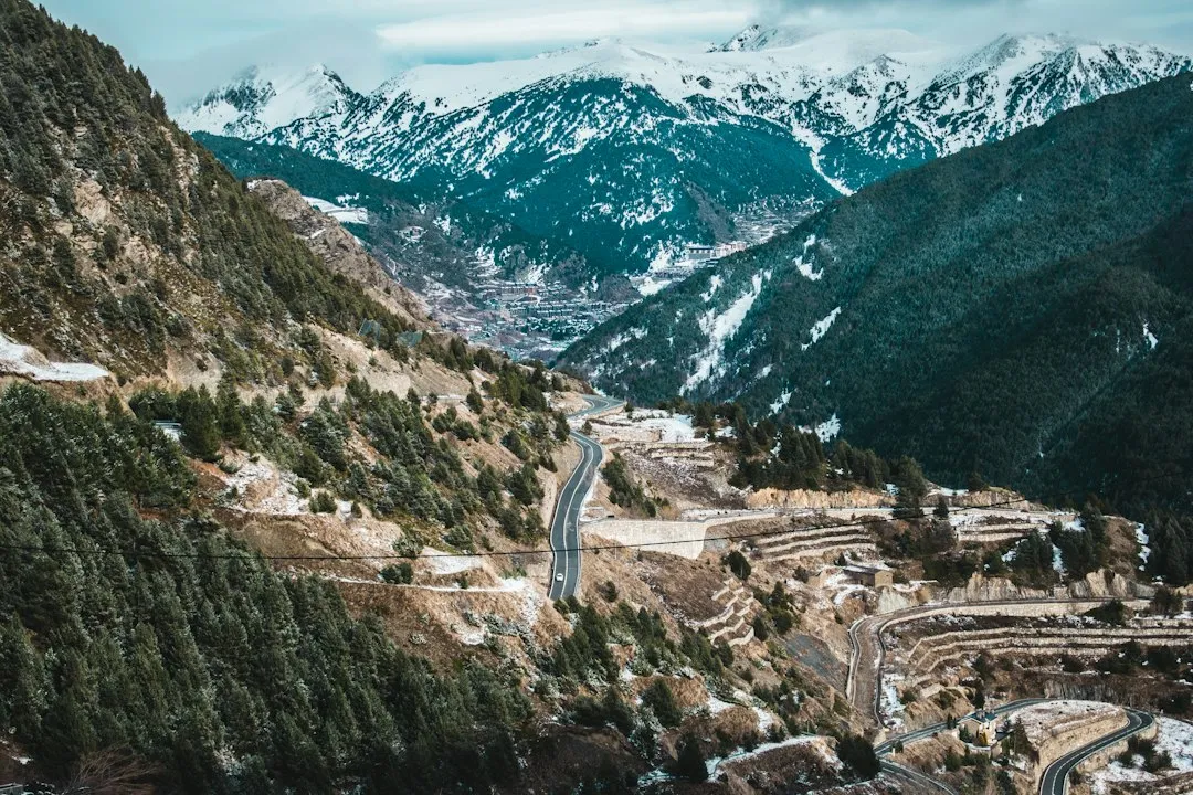 A winding mountain road climbing through a snow-dusted Pyrenean valley in Andorra, with peaks on the horizon.