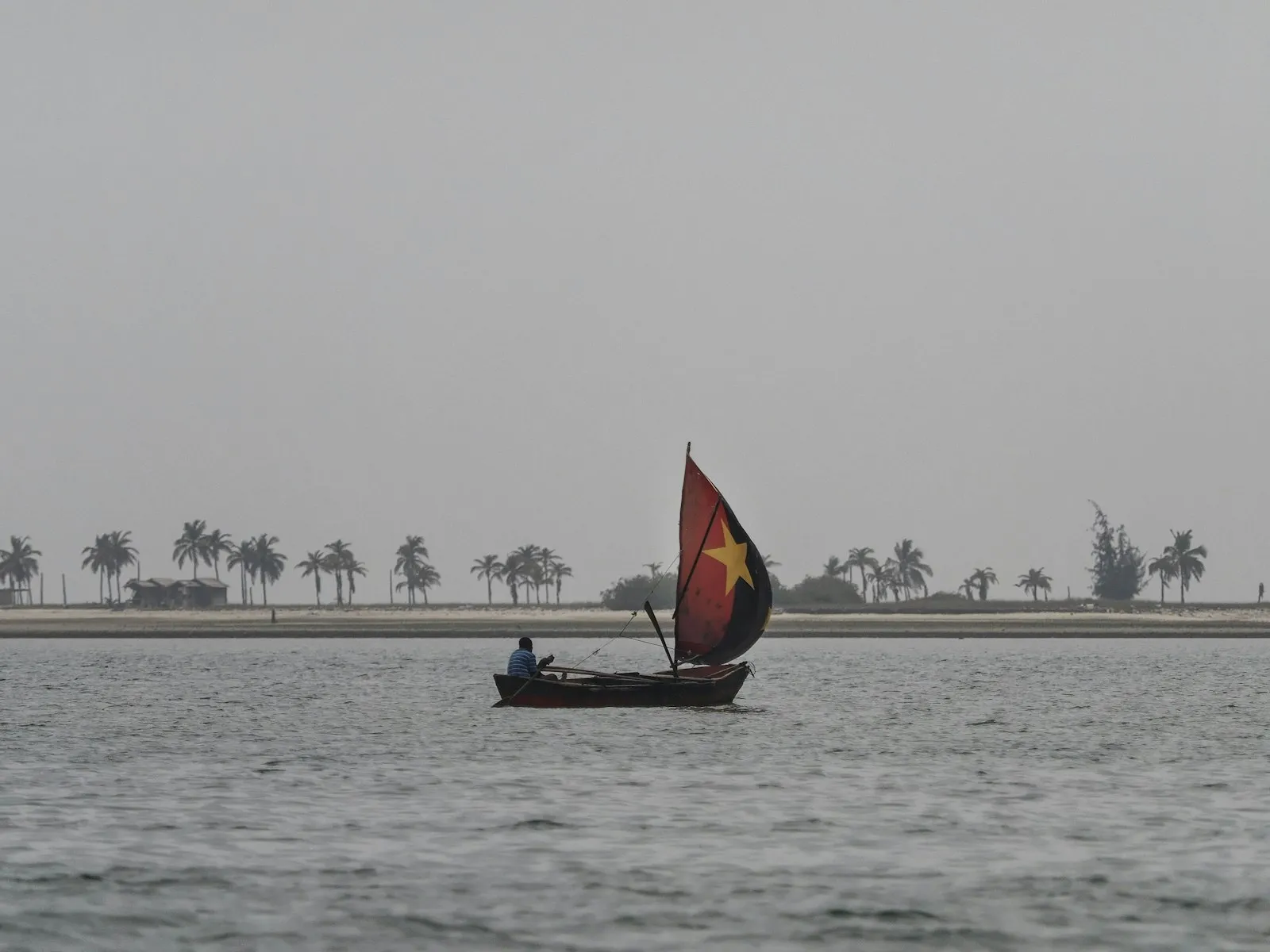 A fisherman's sailboat with a dark red sail on calm grey water, with palm trees along a distant shore on the coast of Angola.