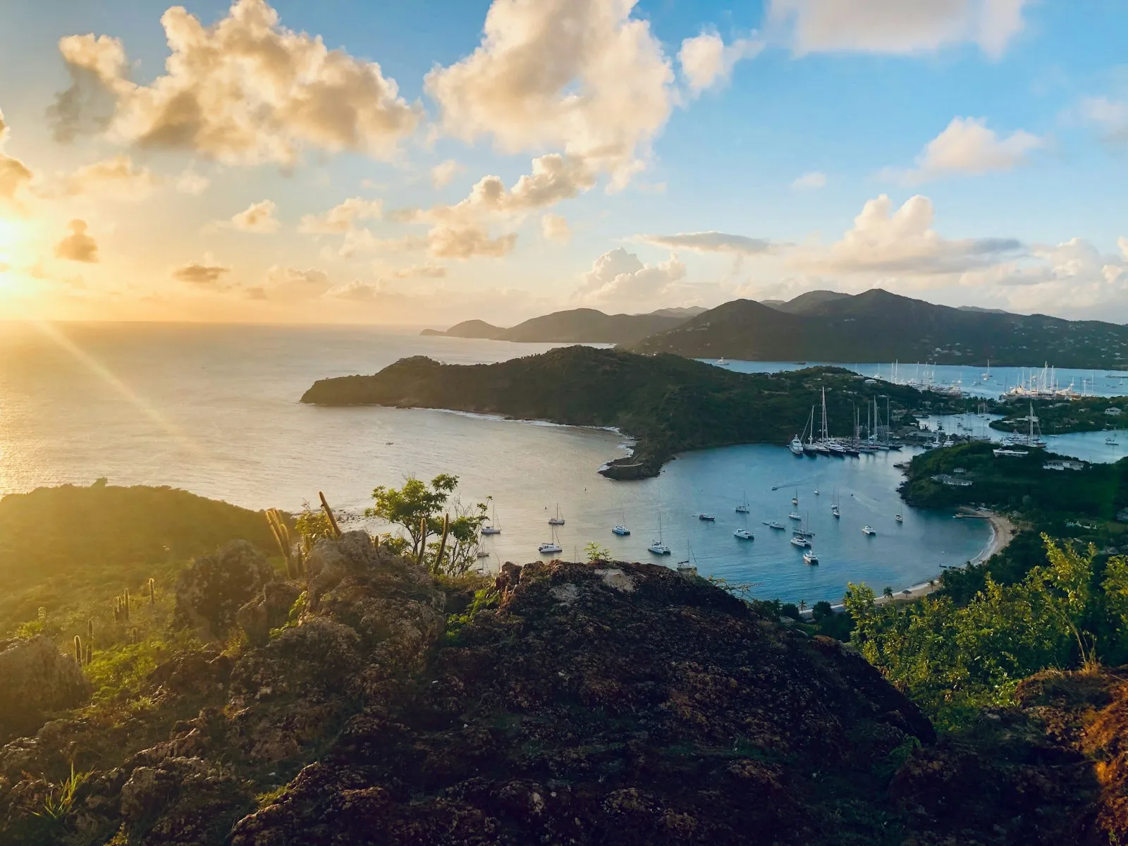 Sailboats at anchor in a wide bay below green hills as the sun sets, seen from a lookout over English Harbour in Antigua.