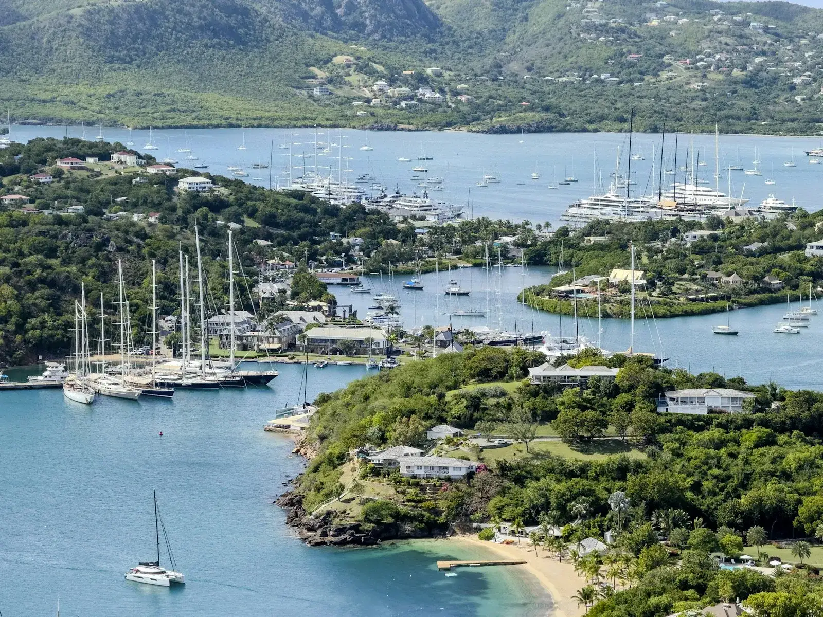 Sailing yachts anchored in a sheltered tropical bay with green hills behind, seen from above on the coast of Antigua.