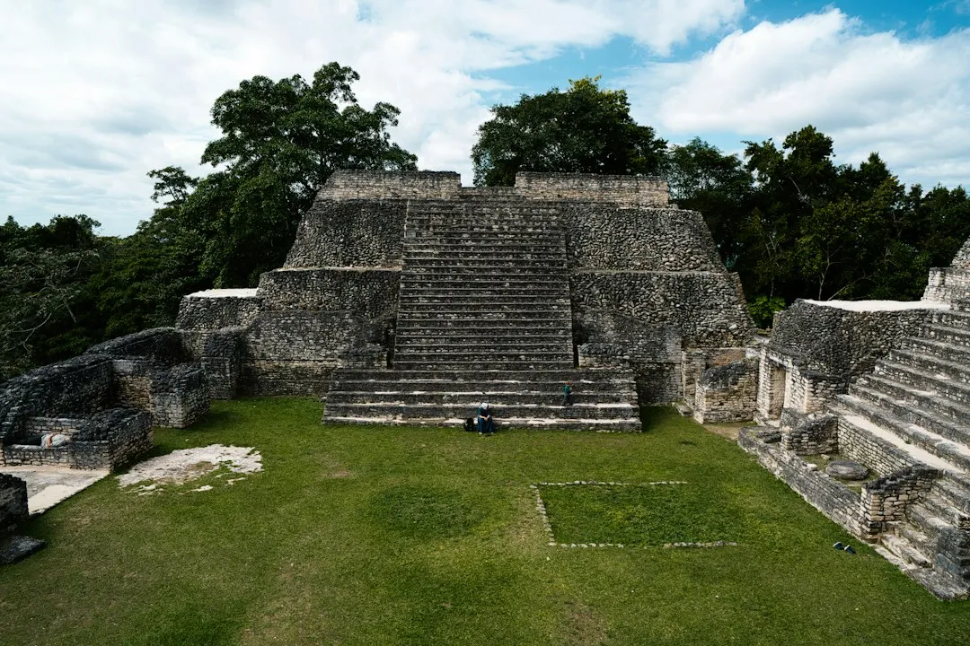 A stone Maya temple with a wide central staircase rising above a mown lawn and tropical trees under a partly cloudy sky in Belize.