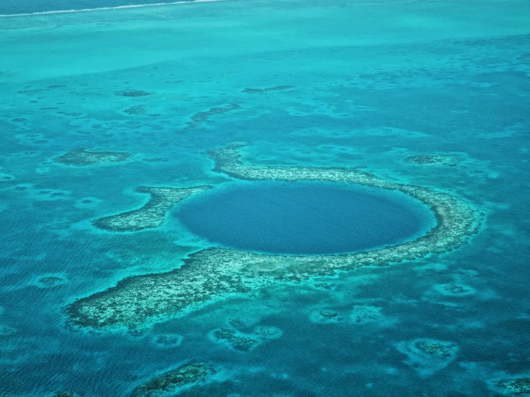 An aerial view of the Great Blue Hole off the coast of Belize: a dark circular sinkhole ringed by pale reef in turquoise water.