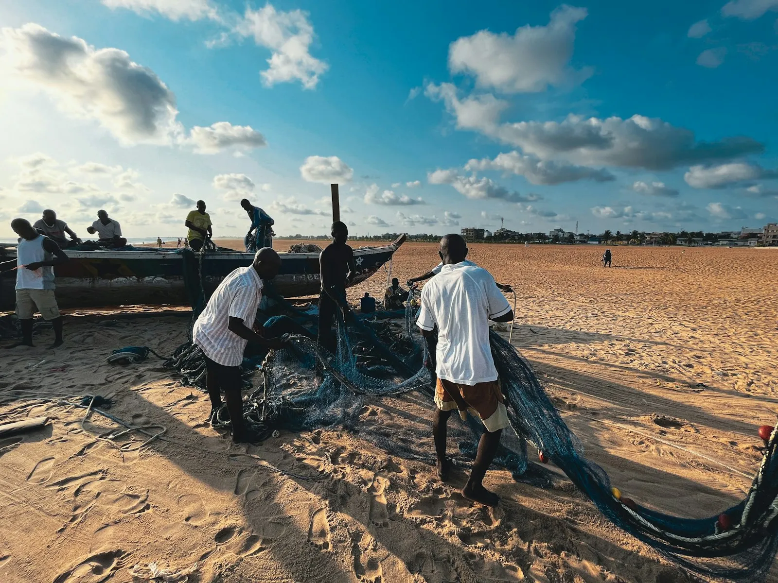 Fishermen working with nets on the sand of Fidjrossè beach in Benin, under a blue sky with scattered clouds.