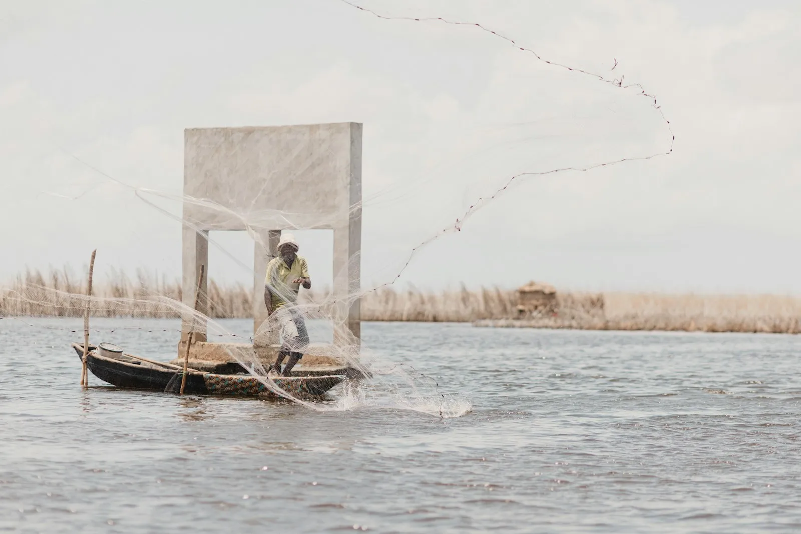 A fisherman casting a net from a wooden canoe on Lake Nokoué near Cotonou, with reeds along the far shore.