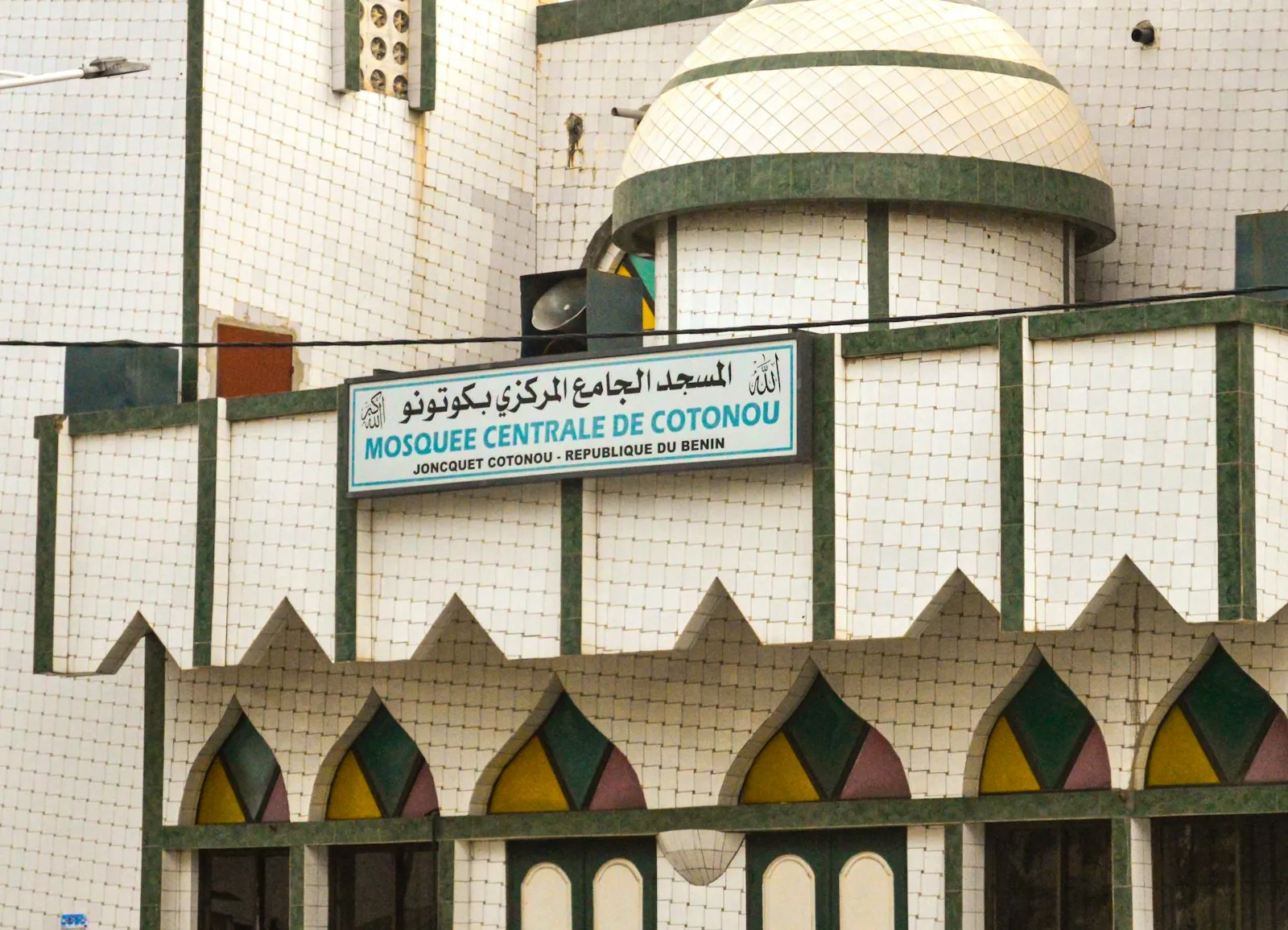 The front of the Central Mosque in Cotonou: a white building with green trim, a dome, arched windows and a sign in Arabic and French.