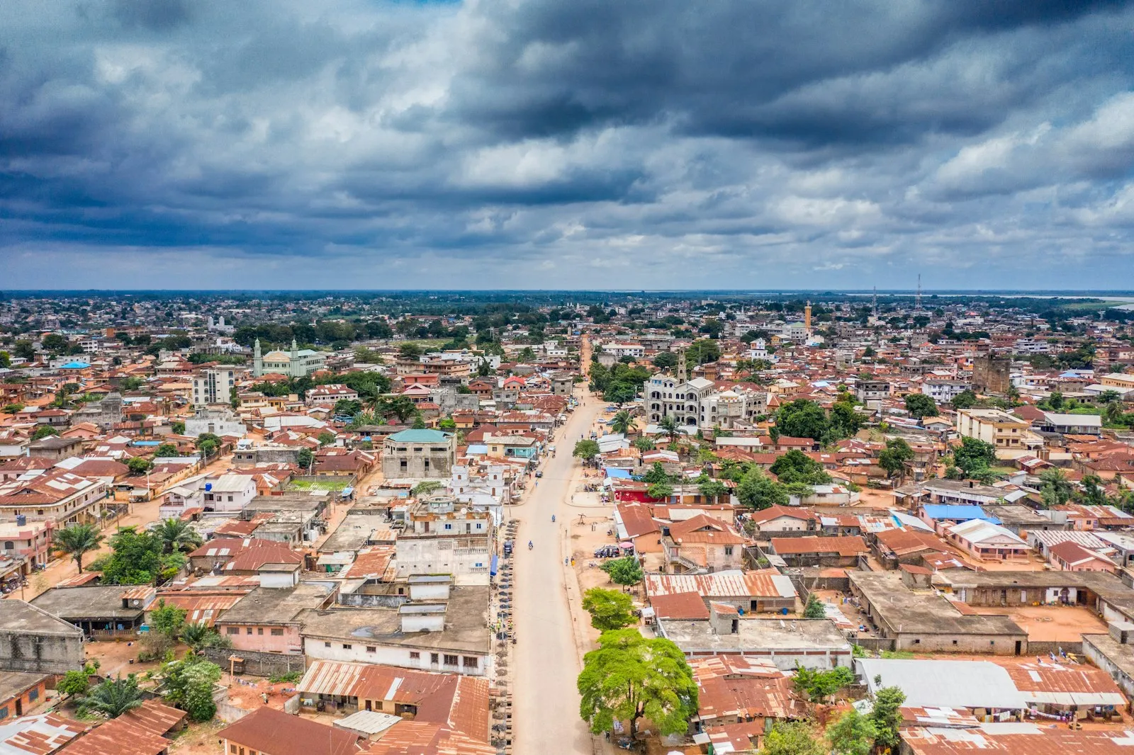 A view over Porto-Novo, the capital of Benin: red-roofed houses and green trees along a straight road, under a heavy cloudy sky.
