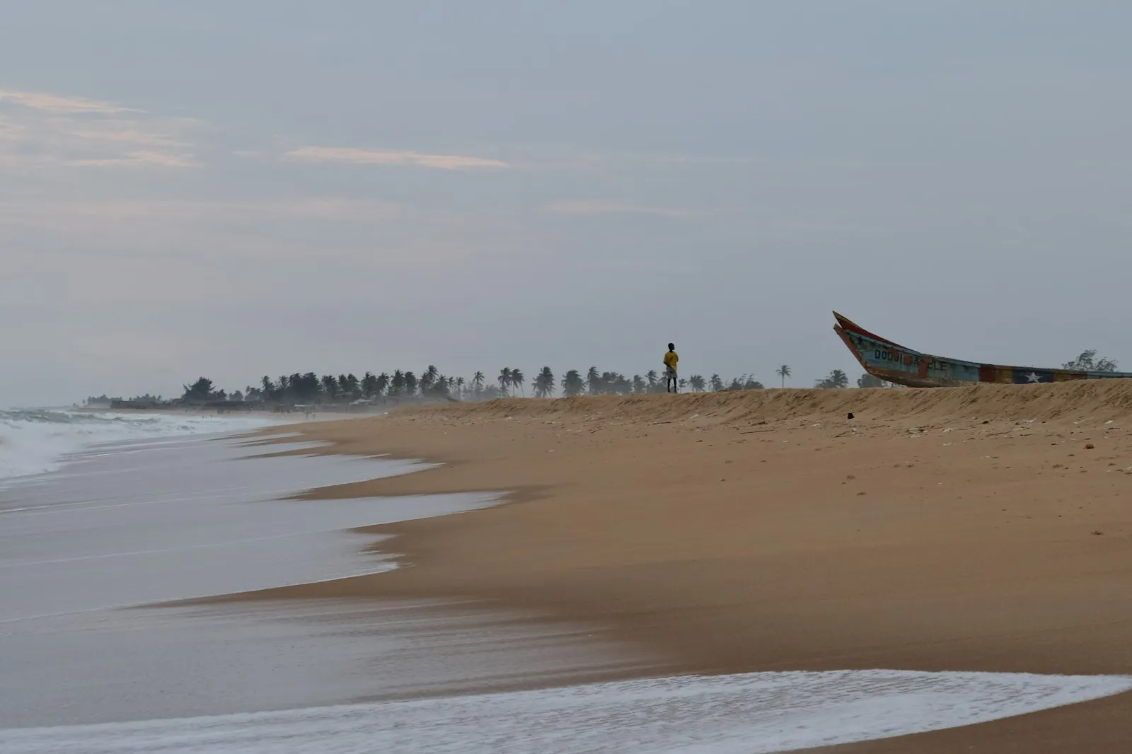 A quiet stretch of beach in Benin with a person standing near a painted fishing boat at the top of the sand, under a pale sky.