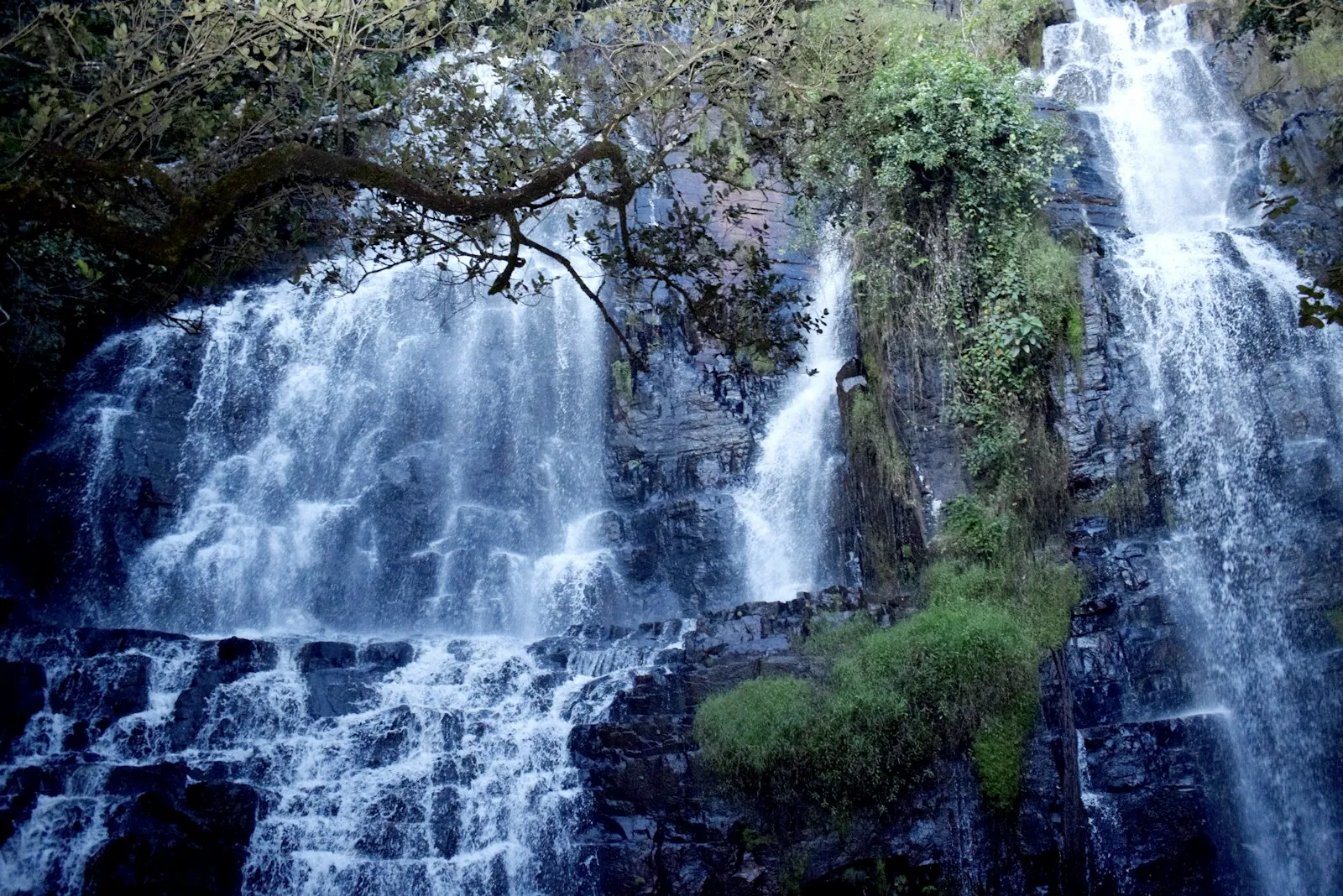 A tall waterfall with several streams falling over dark rock beside green trees in Burundi.