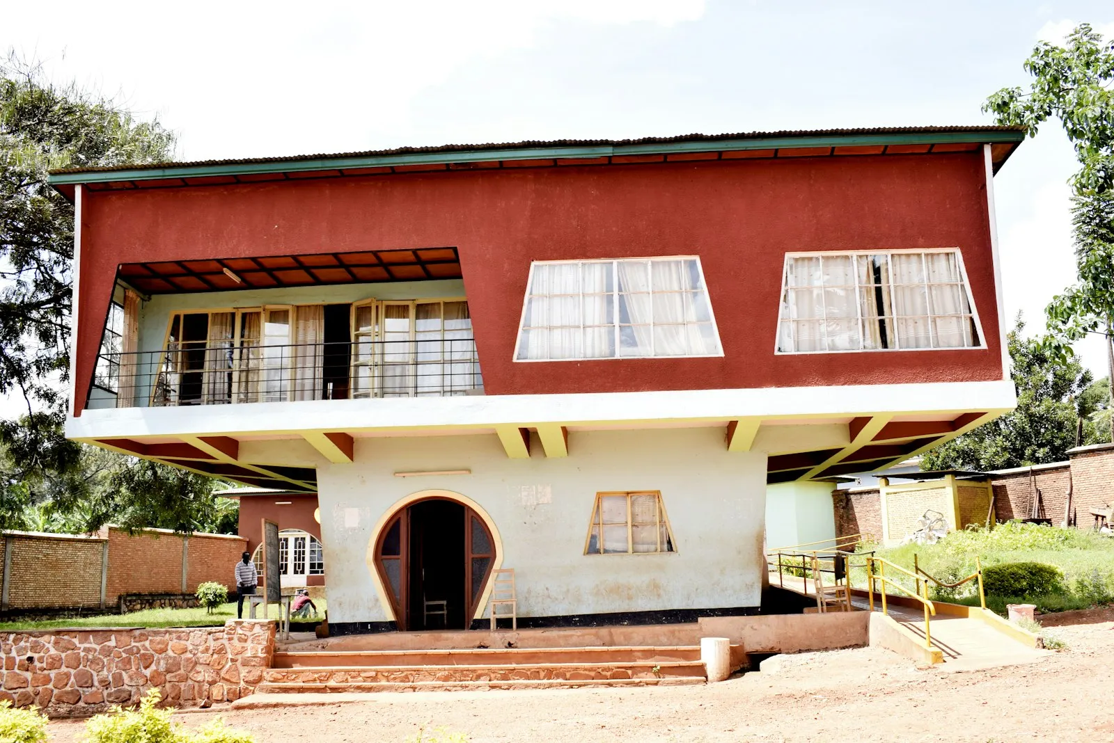 A two-storey building with a red upper floor, a white arched ground floor and a balcony, beside a brick path and trees in Burundi.