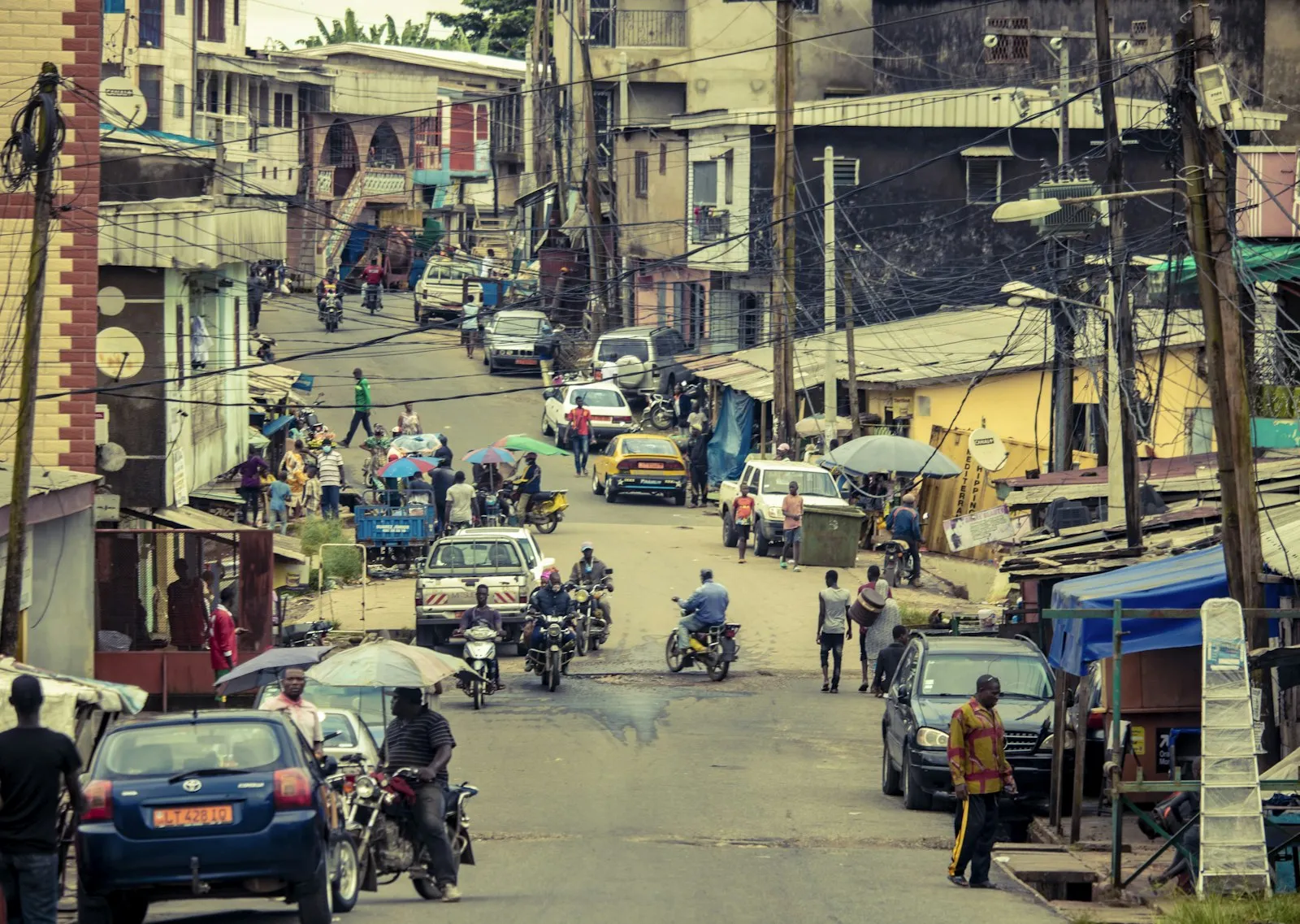 A narrow, crowded street with small shops, motorbikes, cars and pedestrians, lined by tall poles and cables.
