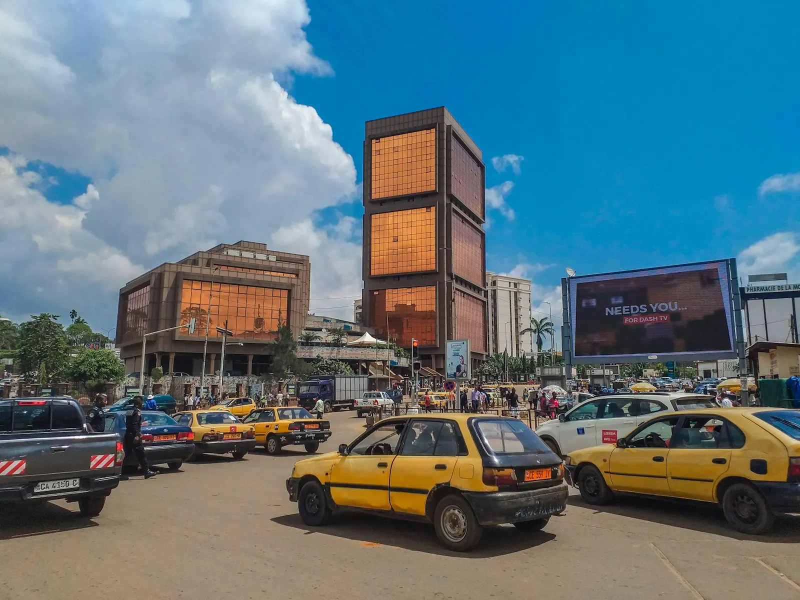A busy city square with yellow taxis in front of two tall buildings with copper-coloured glass facades under a blue sky.
