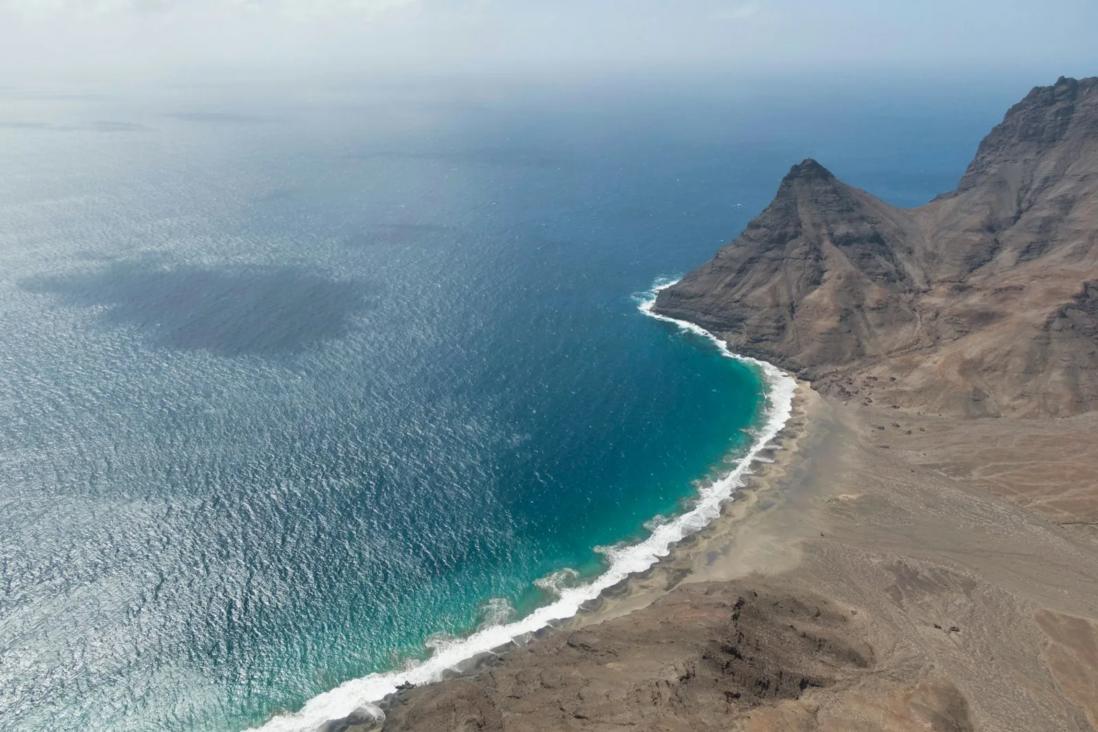 An aerial view of a curved shoreline on the island of S&atilde;o Vicente in Cape Verde: pale beach, white surf and turquoise water below a steep brown headland.