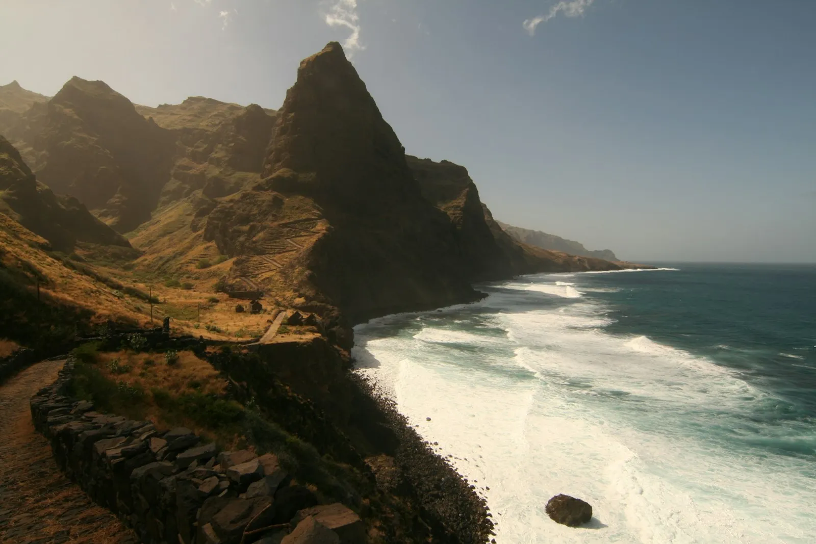 A steep brown mountain coast in Cabo Verde, with a narrow road along a ridge and white surf breaking on the shore below.