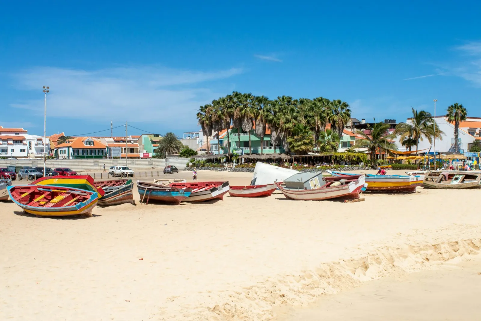 Colourful wooden fishing boats lined up on a sandy beach in Cabo Verde, with palm trees and low buildings behind them under a blue sky.