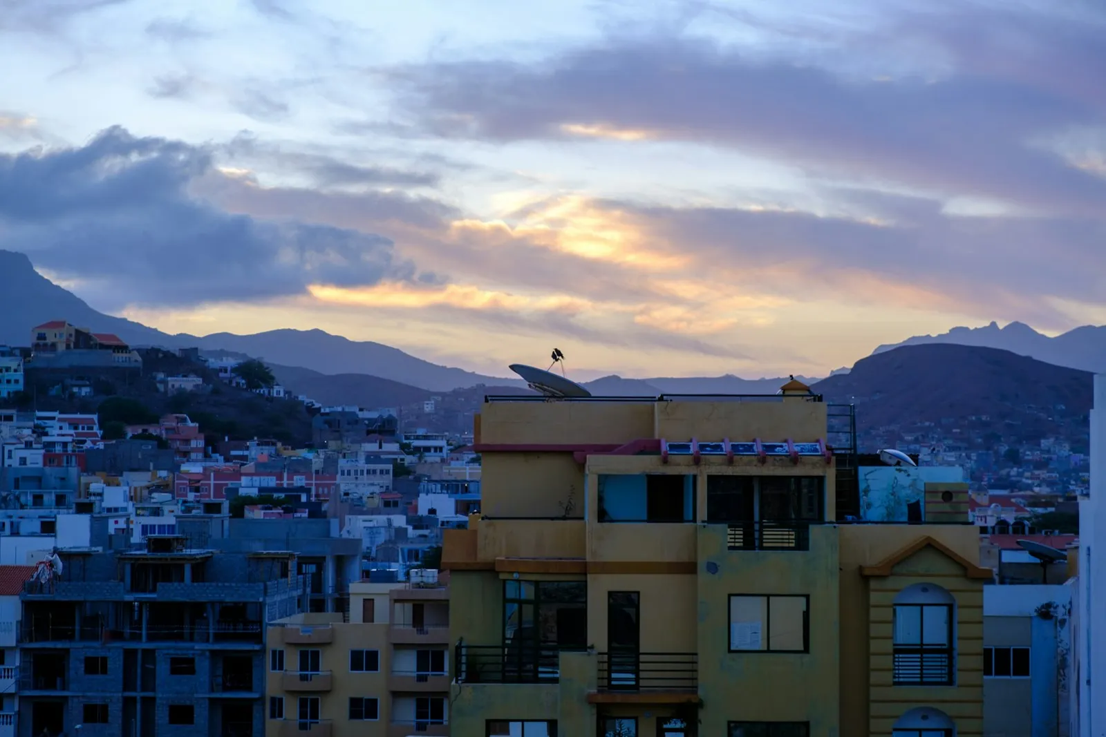 Houses and apartment blocks on a hillside in Mindelo, Cape Verde, with dark mountains behind them under a sunrise sky of pink and grey cloud.