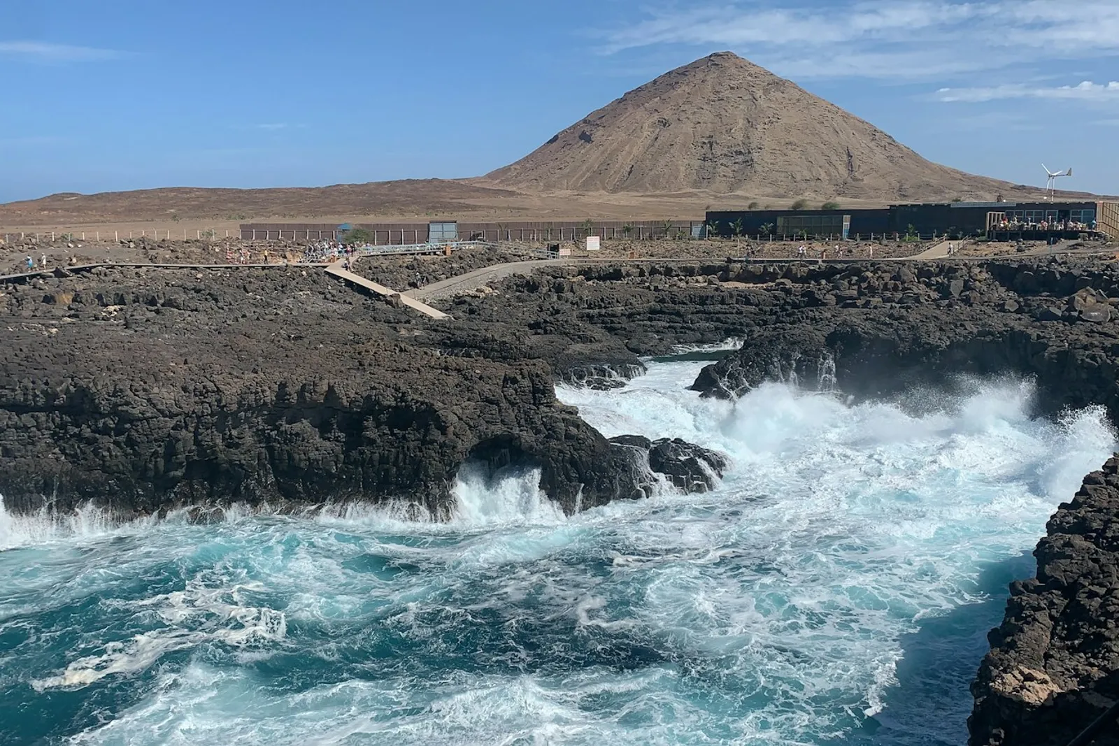 A wave breaking over black volcanic rock beneath a conical brown hill under a pale blue sky on the coast of Cabo Verde.