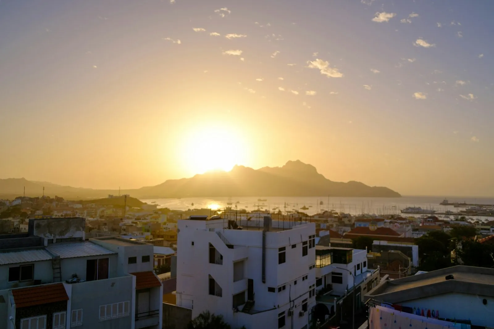 The sun setting behind a dark mountain across the bay at Mindelo, with white houses in the foreground, in Cape Verde.