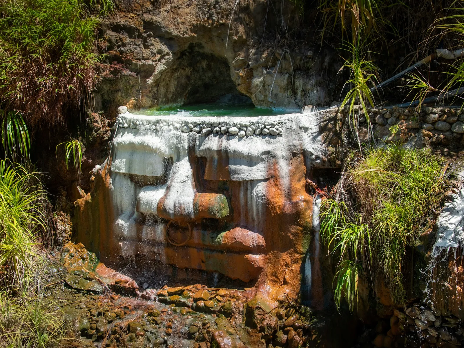 A small waterfall pouring over orange-stained volcanic rock into a hot spring pool at Titou Gorge, ringed by tropical plants.