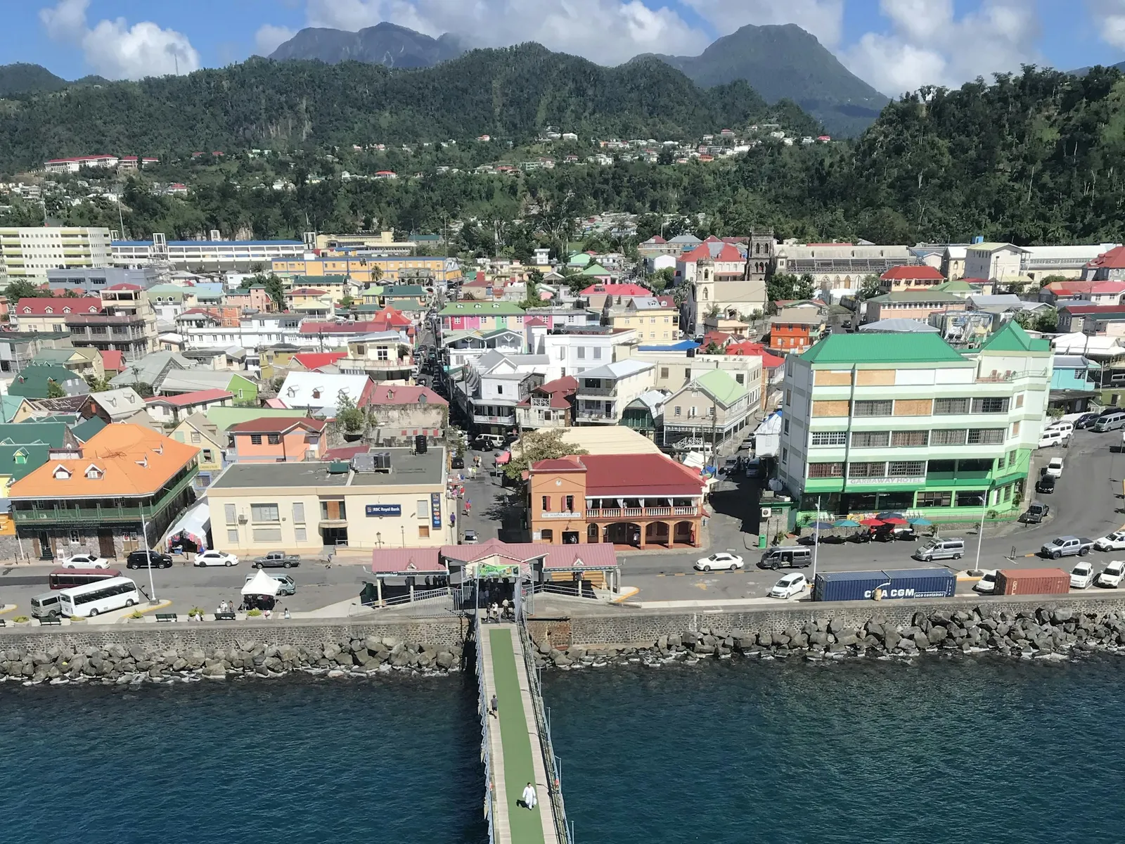 An aerial view of Roseau's waterfront with a long pier and the town's coloured buildings below green mountains.
