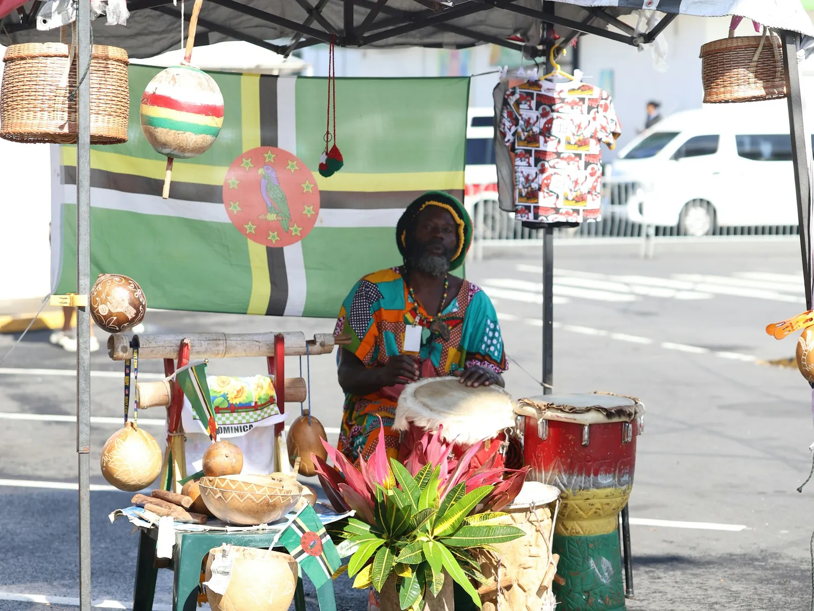 A craft seller in a bright shirt behind a market stall hung with a Dominica flag, drums and calabash gourds.