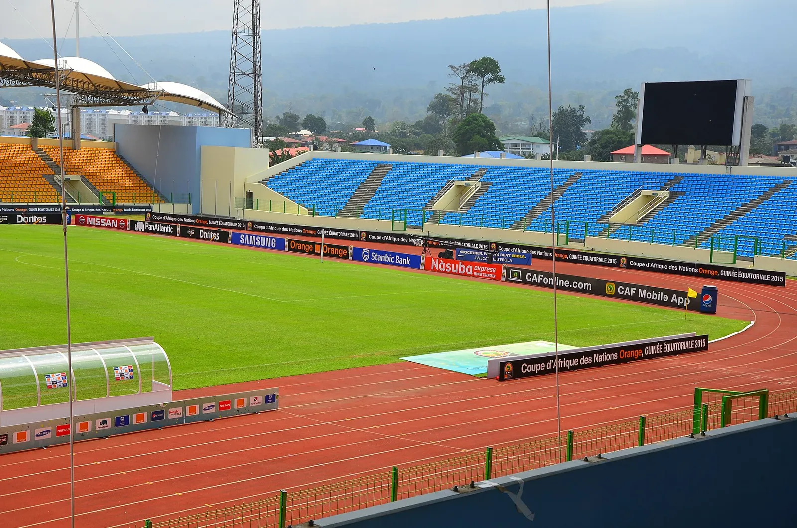 A modern stadium with blue and orange seating, a running track and a green pitch, with hills behind it under a hazy sky, in Malabo, Equatorial Guinea.