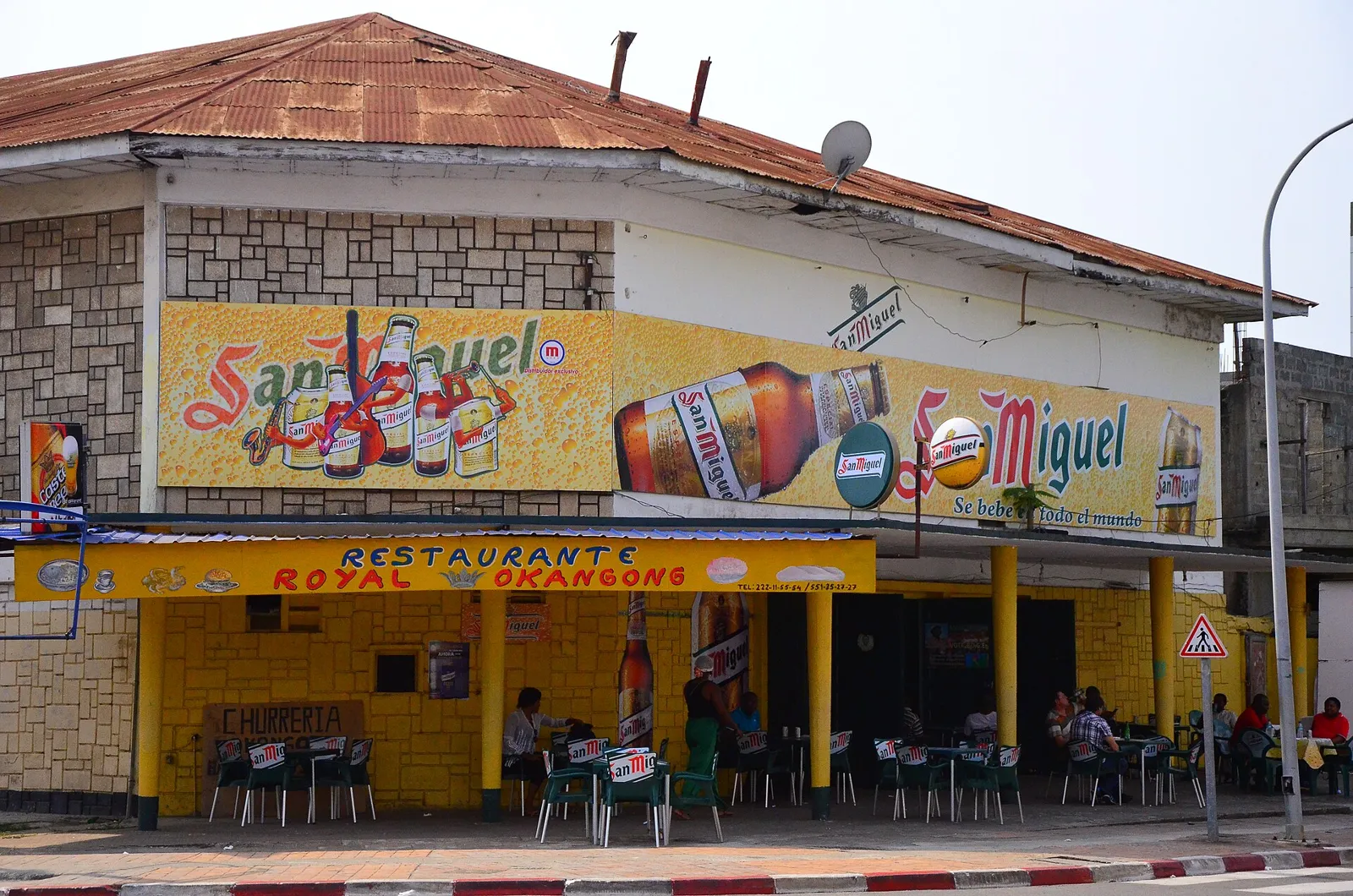 A yellow and red restaurant building on a street corner in Bata, with a San Miguel sign across its front and people sitting under the awning.