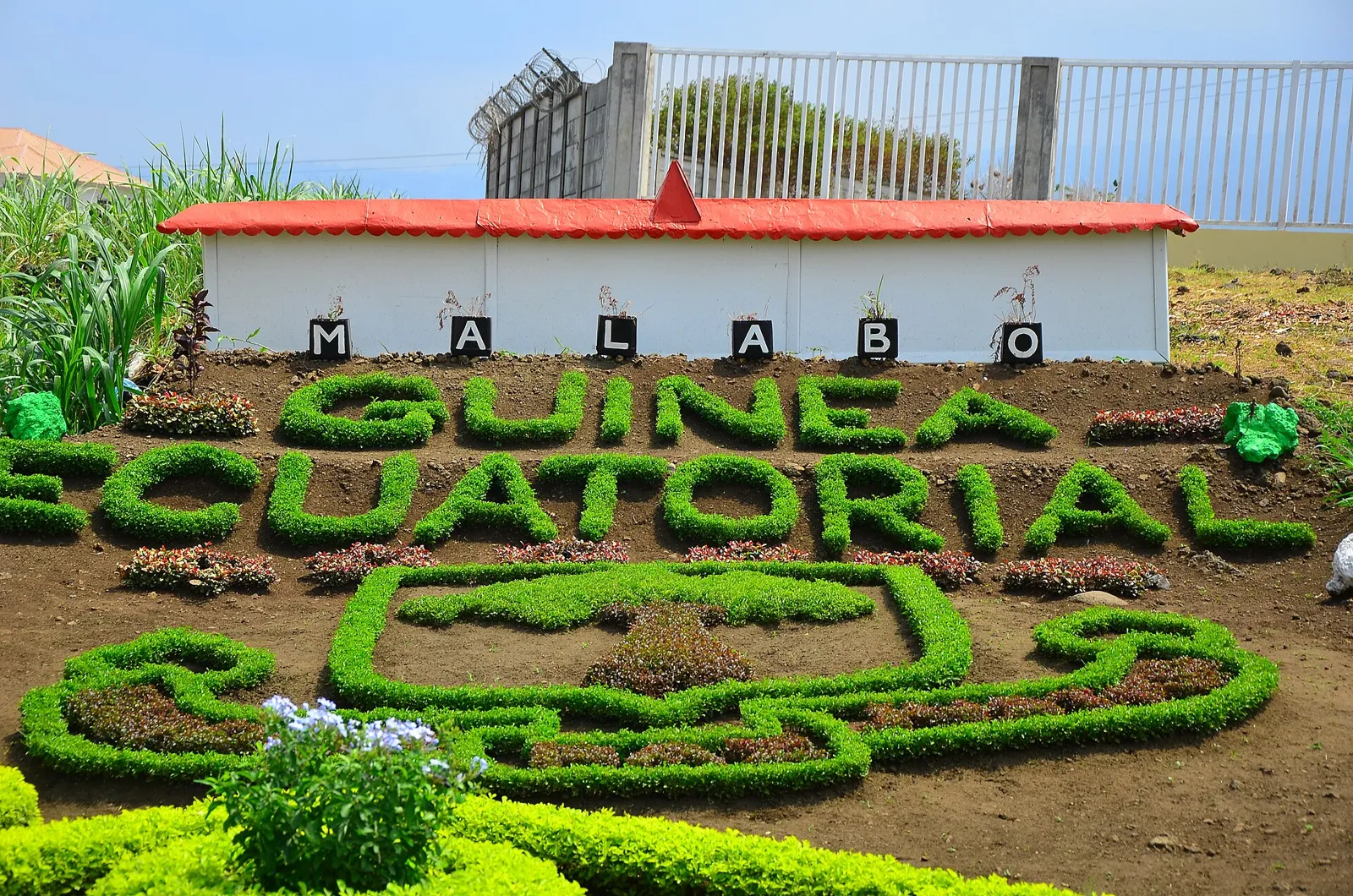 A garden bed of clipped green plants spelling out Guinea Ecuatorial, with a Malabo sign above it and a low building behind.