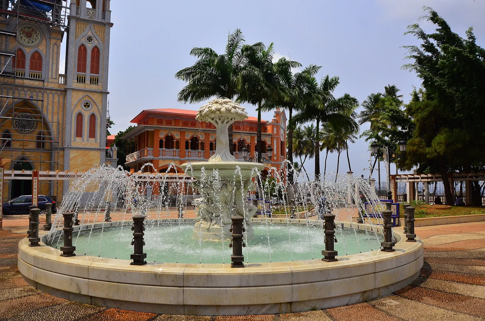 A fountain with palm trees in a sunlit square, with a twin-towered church and a pink building behind it, in Malabo, Equatorial Guinea.