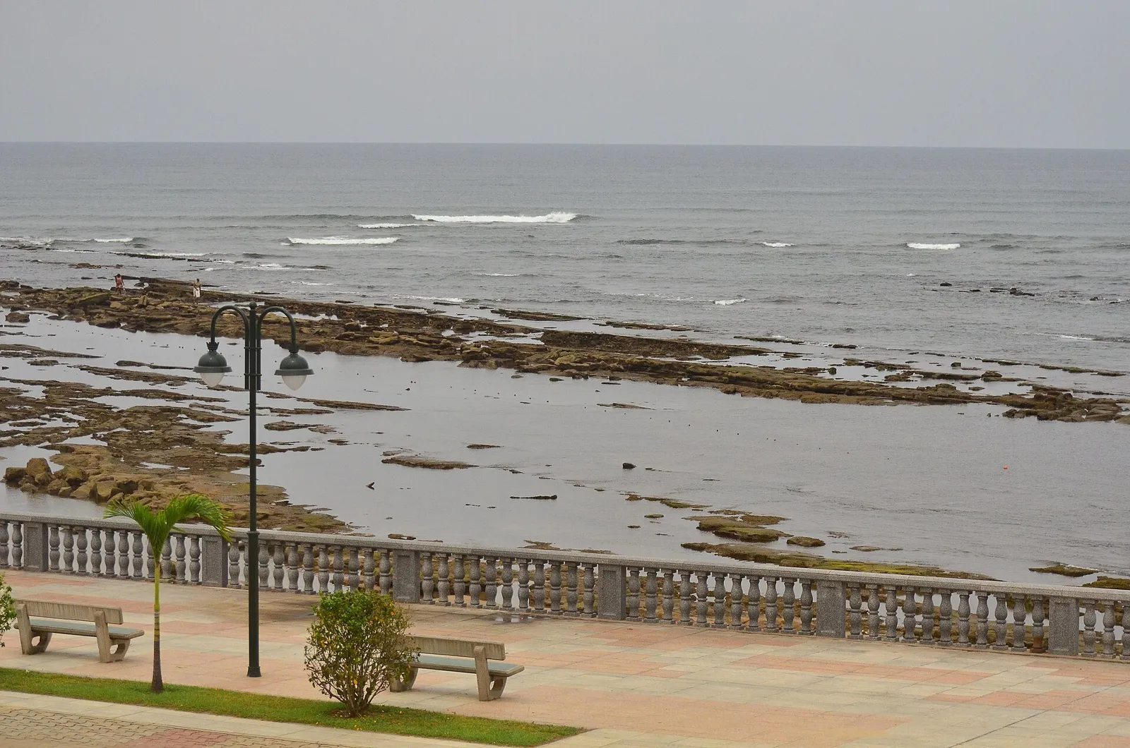 A stone-balustraded seafront promenade with lamp posts and benches above a wide, rocky shore, with grey surf and sky beyond.