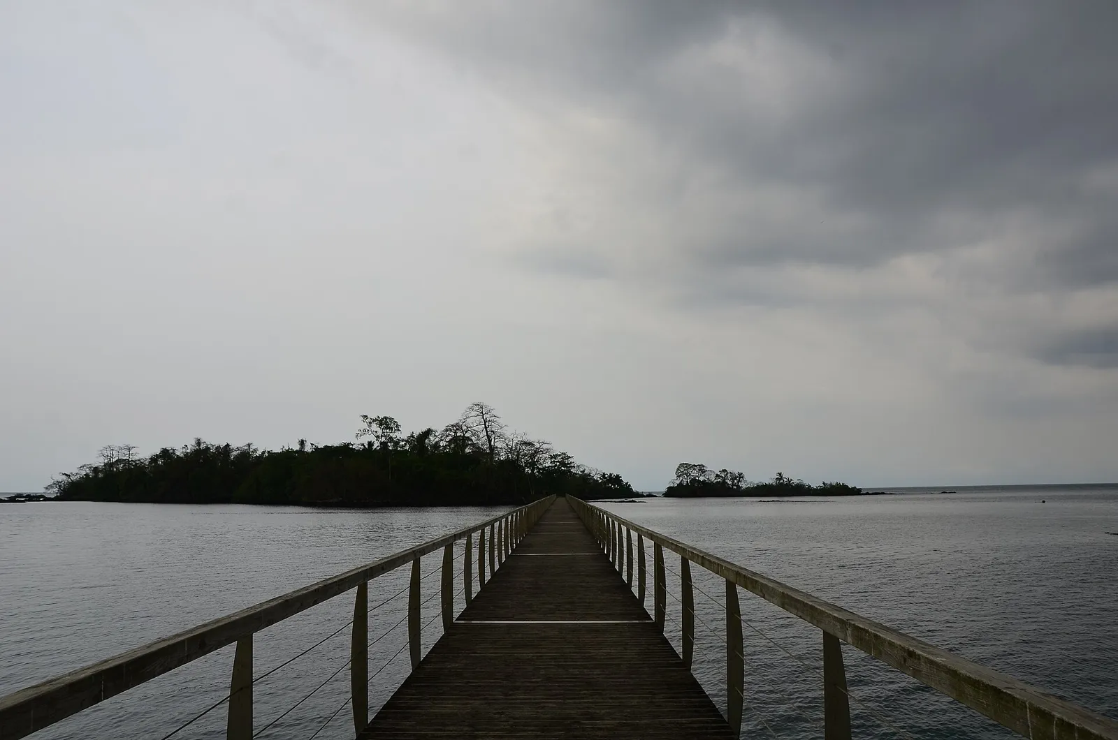 A long wooden jetty running out over still grey water towards a small tree-covered island under a cloudy sky.
