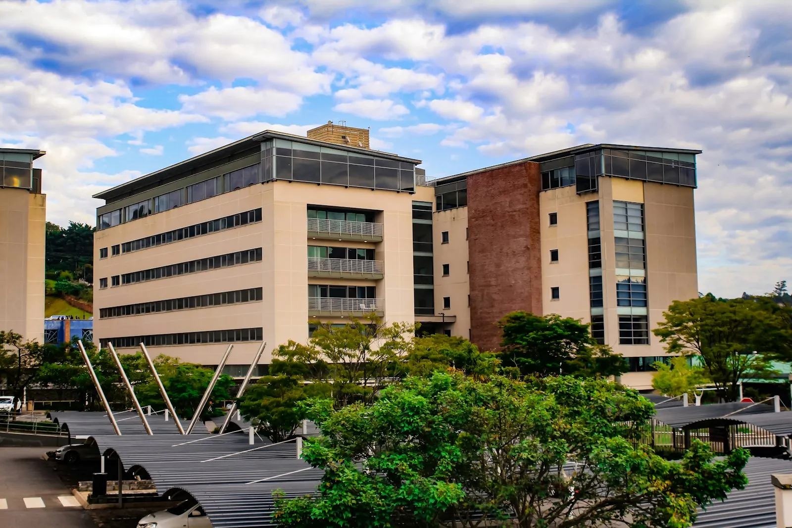 A modern office building with glass and tiled walls, trees in front and a cloudy blue sky behind.