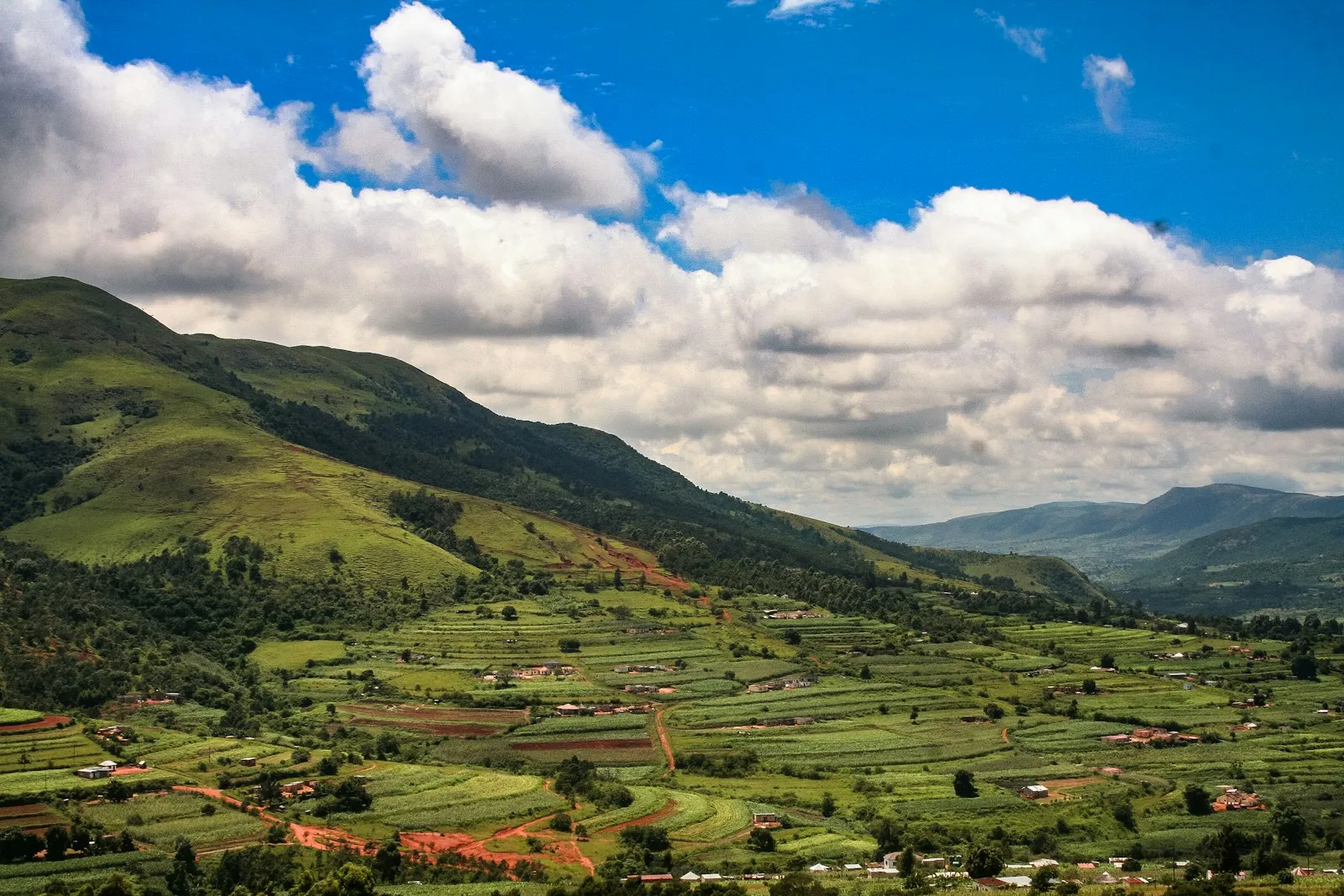 Terraced green fields in a valley below steep green hills, under blue sky and large white clouds.