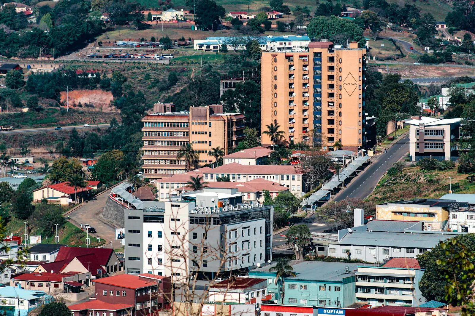 Office blocks, a tall tower and red-roofed buildings climbing a green hillside in Mbabane, with hills rising behind.