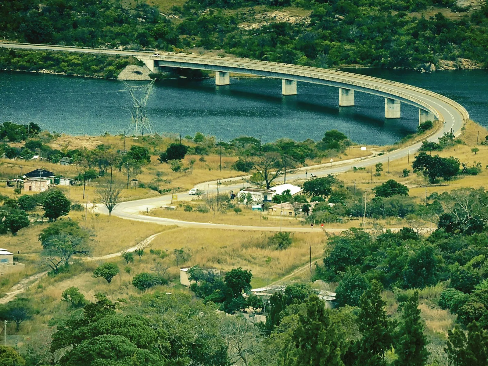 A curving road bridge crossing calm blue water between green and golden land.