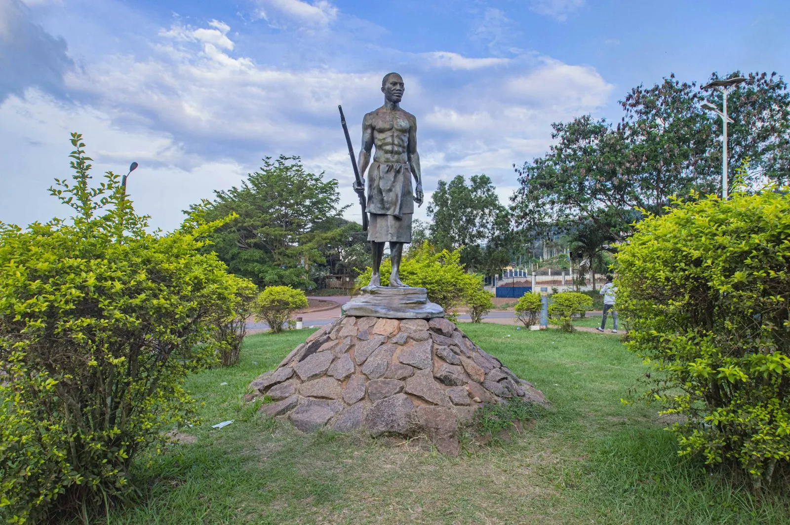 A bronze statue of a standing warrior with a spear on a rocky plinth in a garden at Lastoursville, Gabon, under a blue sky.