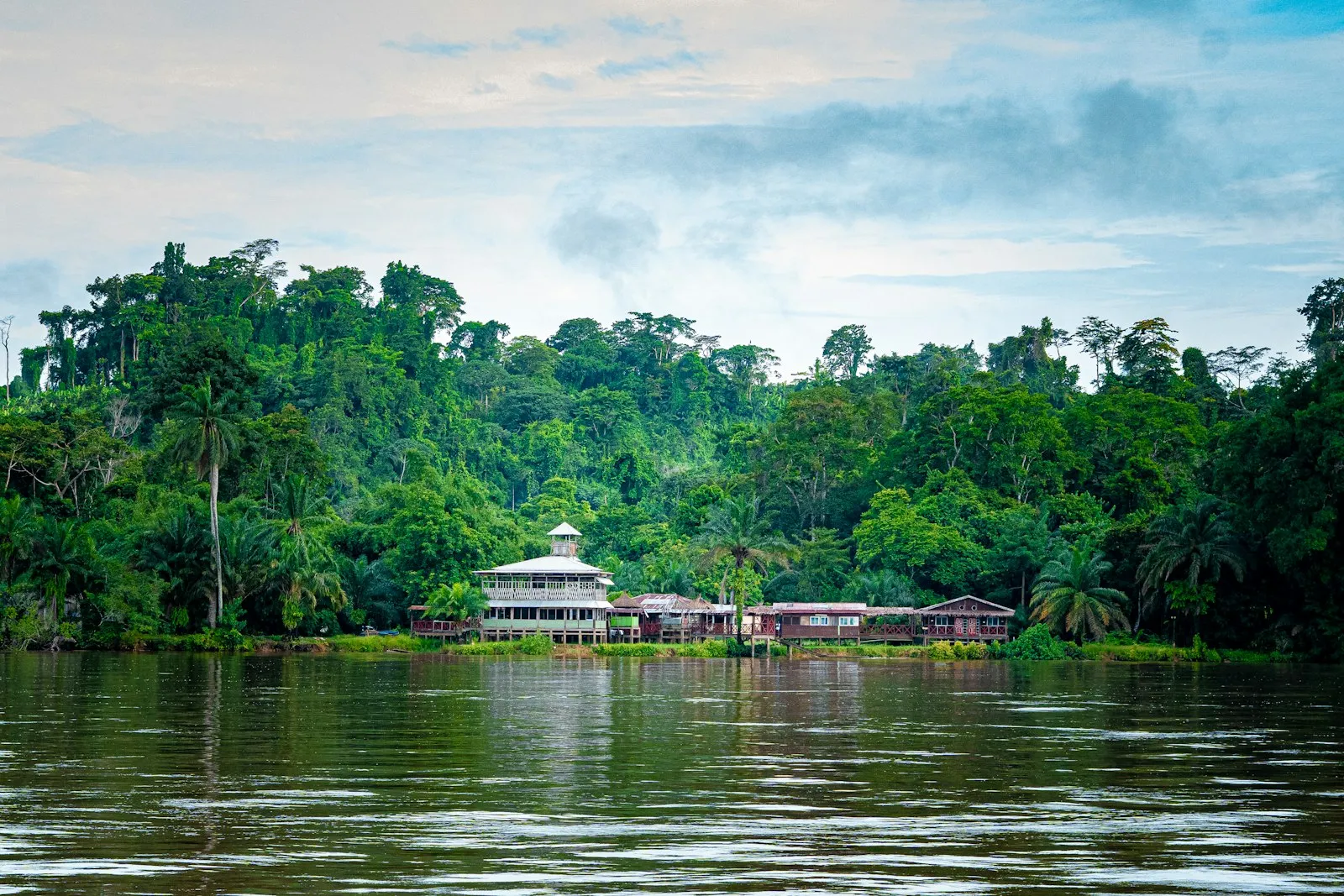 Houses and a white building with a tower beside the Ogooué river at Lambaréné, Gabon, set among dense green forest.