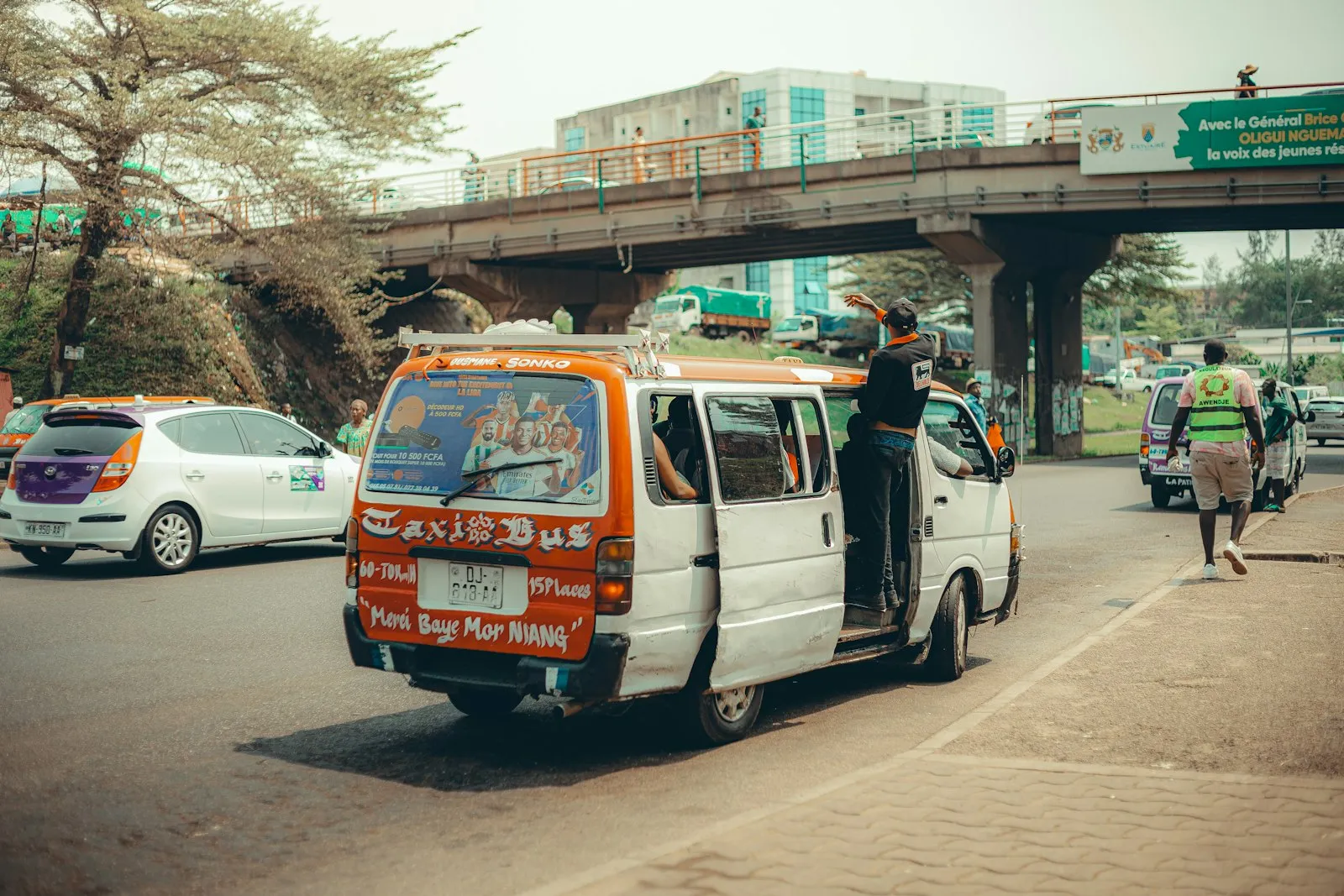 A white minibus taxi with its door open on a busy road in Libreville, Gabon, with cars, a flyover and buildings behind.