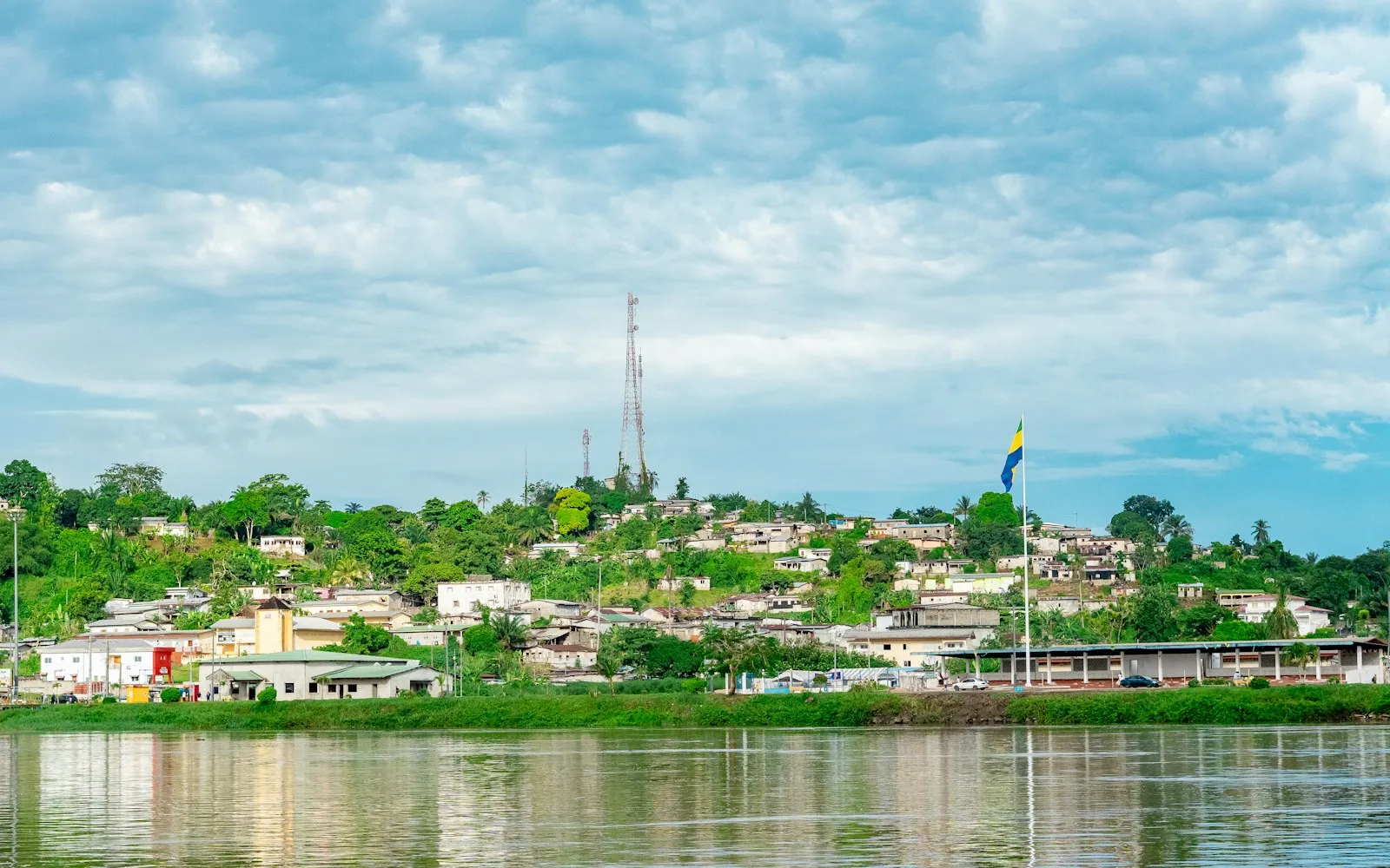 Houses and a hilltop radio mast on the bank of the Ogooué river at Lambaréné, Gabon, under a blue sky with scattered cloud.