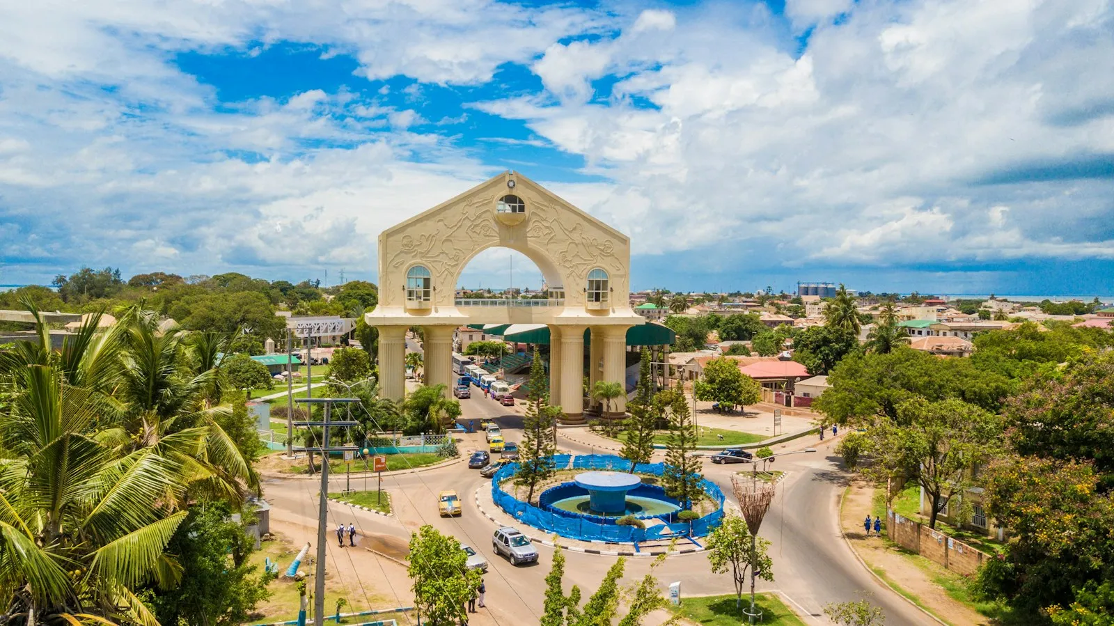 Arch 22 in Banjul, a tall pale stone arch beside a roundabout fountain, with palm trees and low buildings under a blue sky with white clouds.