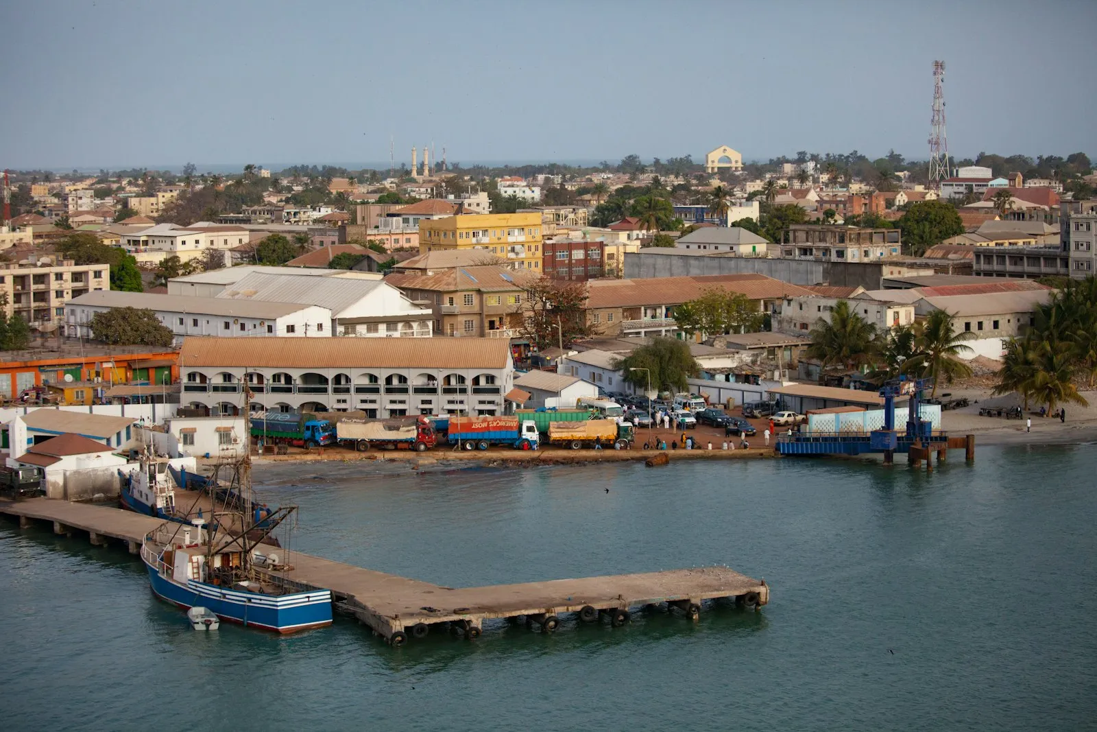 The harbour at Banjul seen from above, with a wooden pier, boats, trucks on the quay and low town buildings behind.