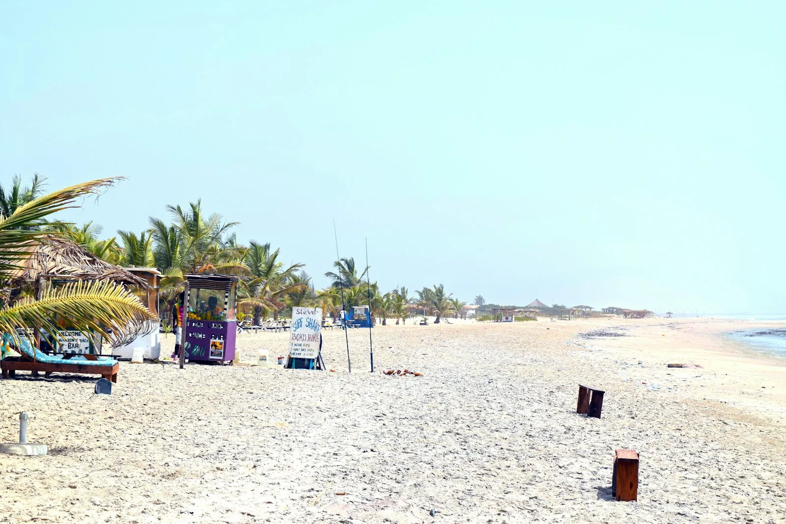 A pale sandy beach with thatched huts, a small kiosk and palm trees under a clear sky.