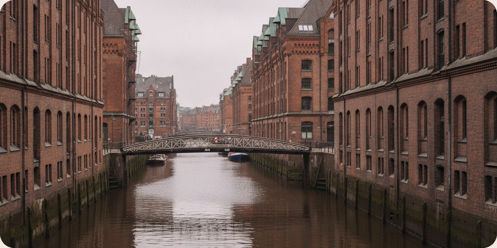 Red-brick warehouse buildings along a canal in Hamburg's Speicherstadt, with a small iron footbridge under a soft overcast sky.