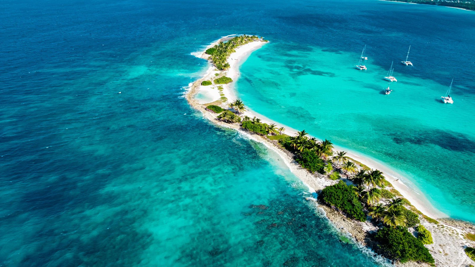 A long sandy spit with palm trees and sailing boats at anchor in turquoise water off the coast of Grenada, seen from above.