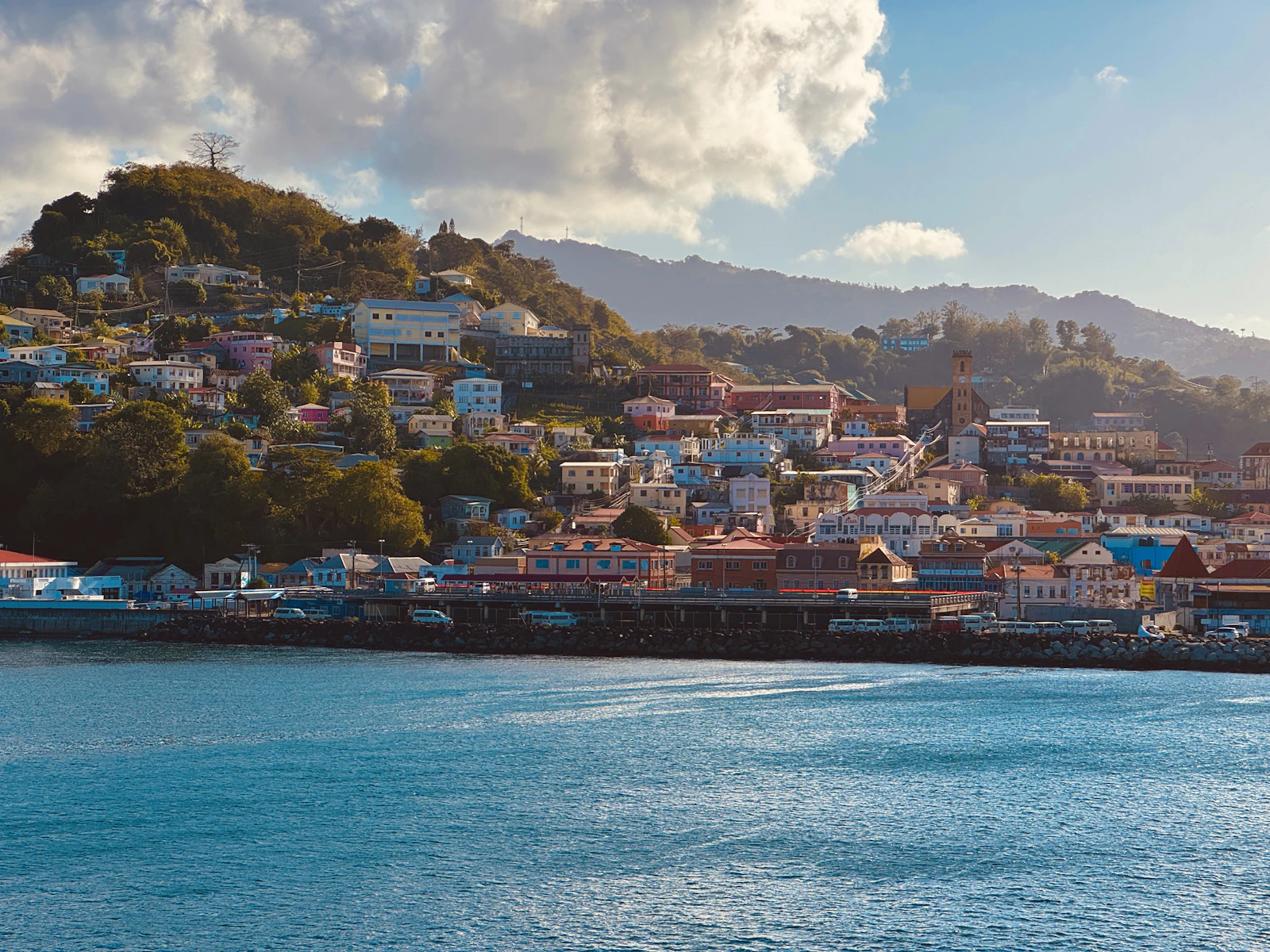Colourful houses and a church steeple climbing a green hill above a calm blue harbour in St. George's, Grenada.