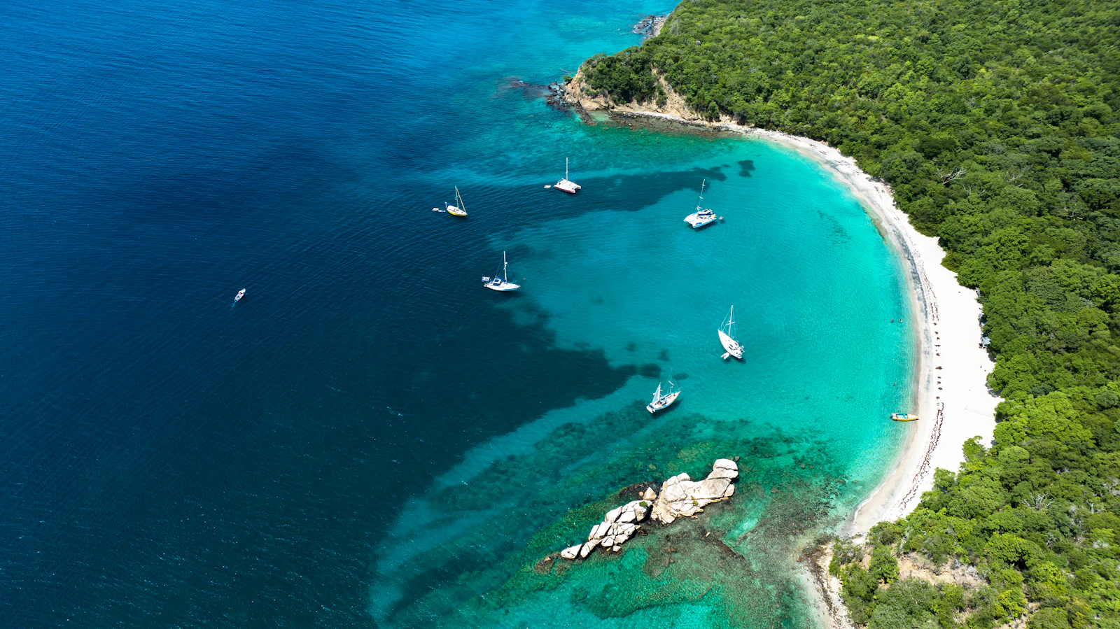 A curved white beach and a cluster of sailing boats in clear turquoise water at Anse La Roche on Carriacou, Grenada.