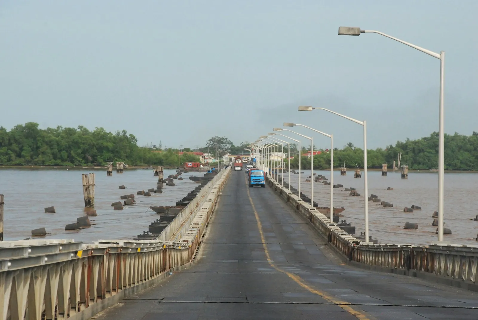 A long road bridge with street lights crossing a wide brown river, with forest on the far bank, in Guyana.