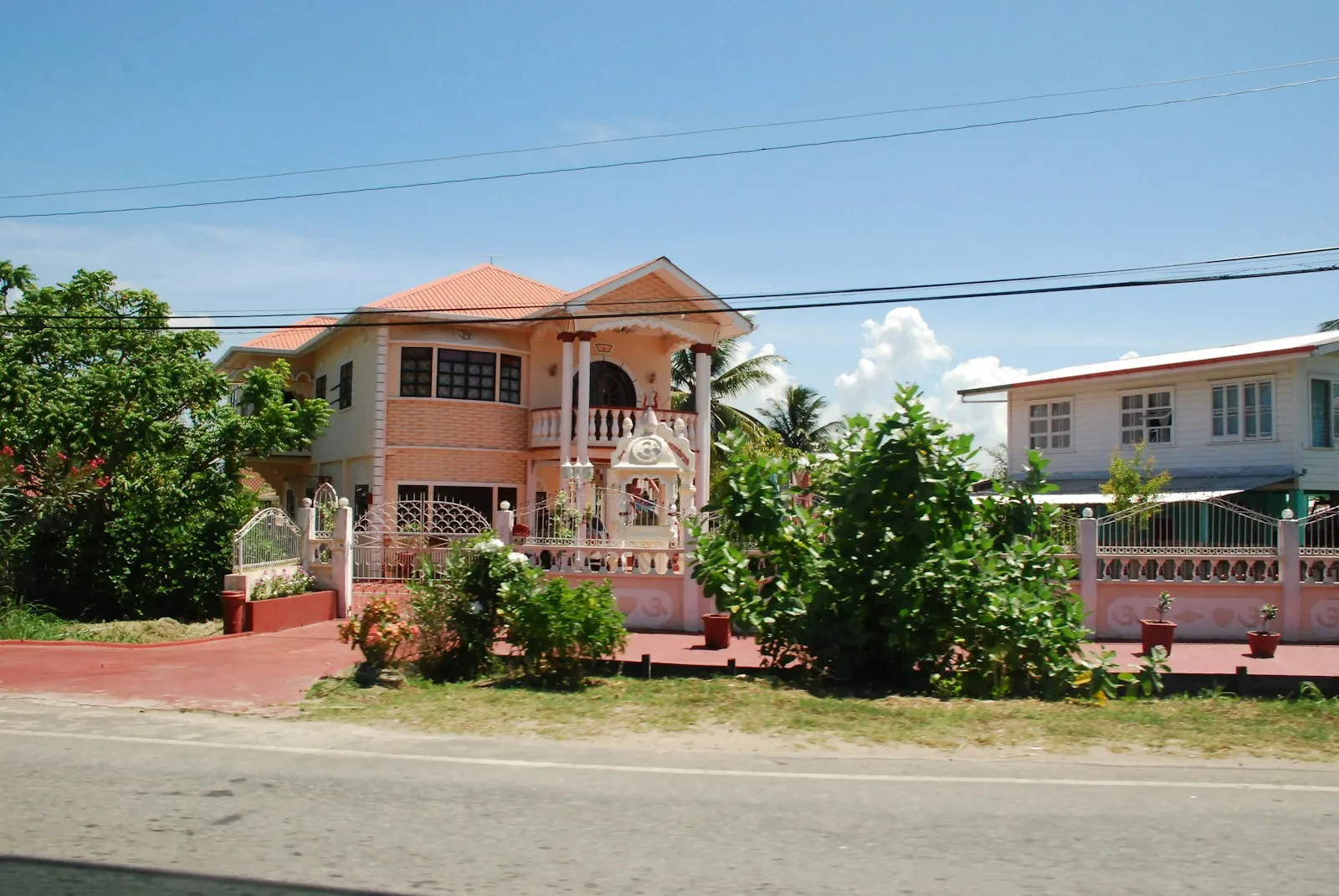 A pink and cream two-storey house with a gate and palm trees beside a road under a blue sky in Guyana.