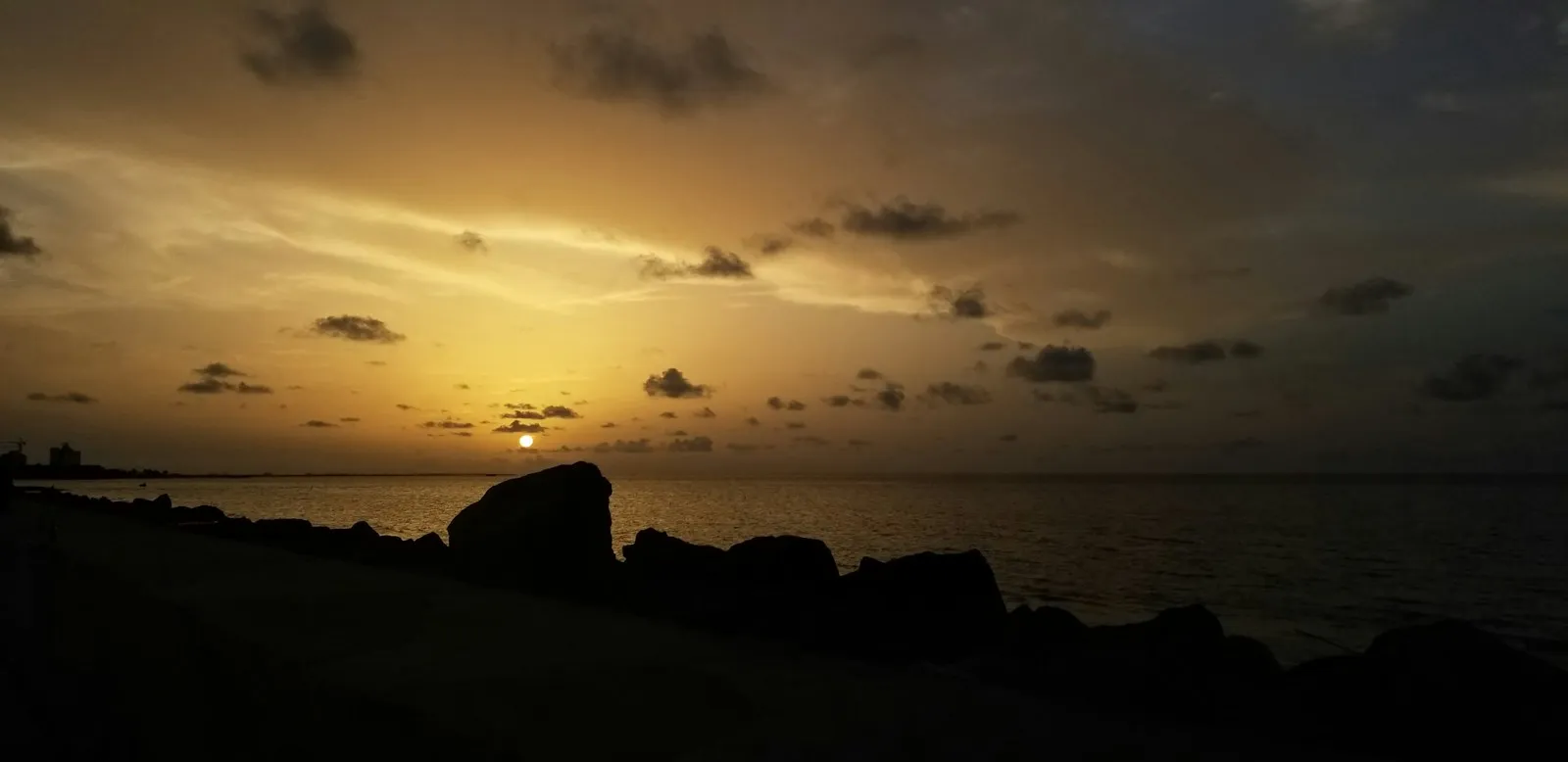 Dark rocks on the seawall in Georgetown with the sun low over a calm sea at dusk.